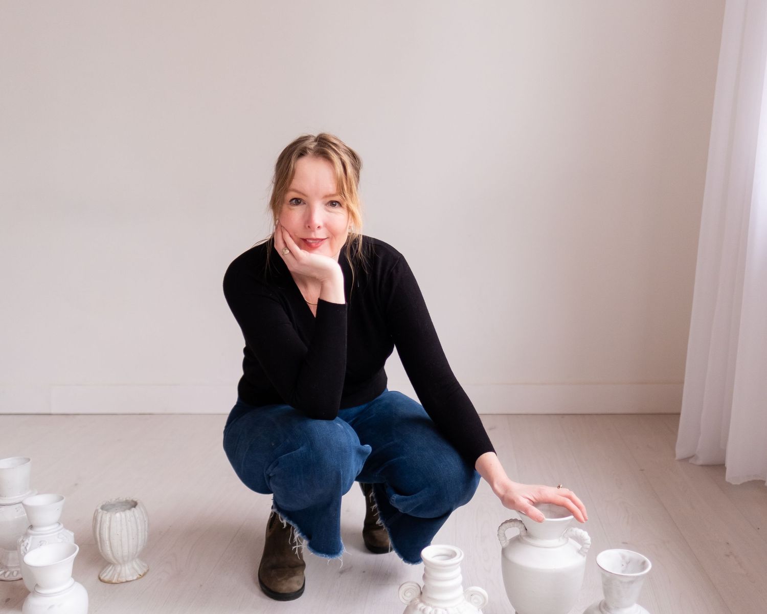 Woman crouching near white ceramic vessels on a light wooden floor in a bright minimalist room.