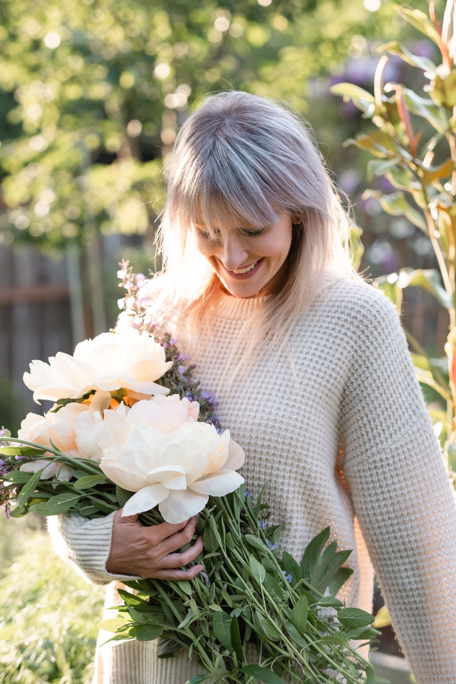 Smiling woman in beige sweater holding large bouquet of white peonies in sunny garden.