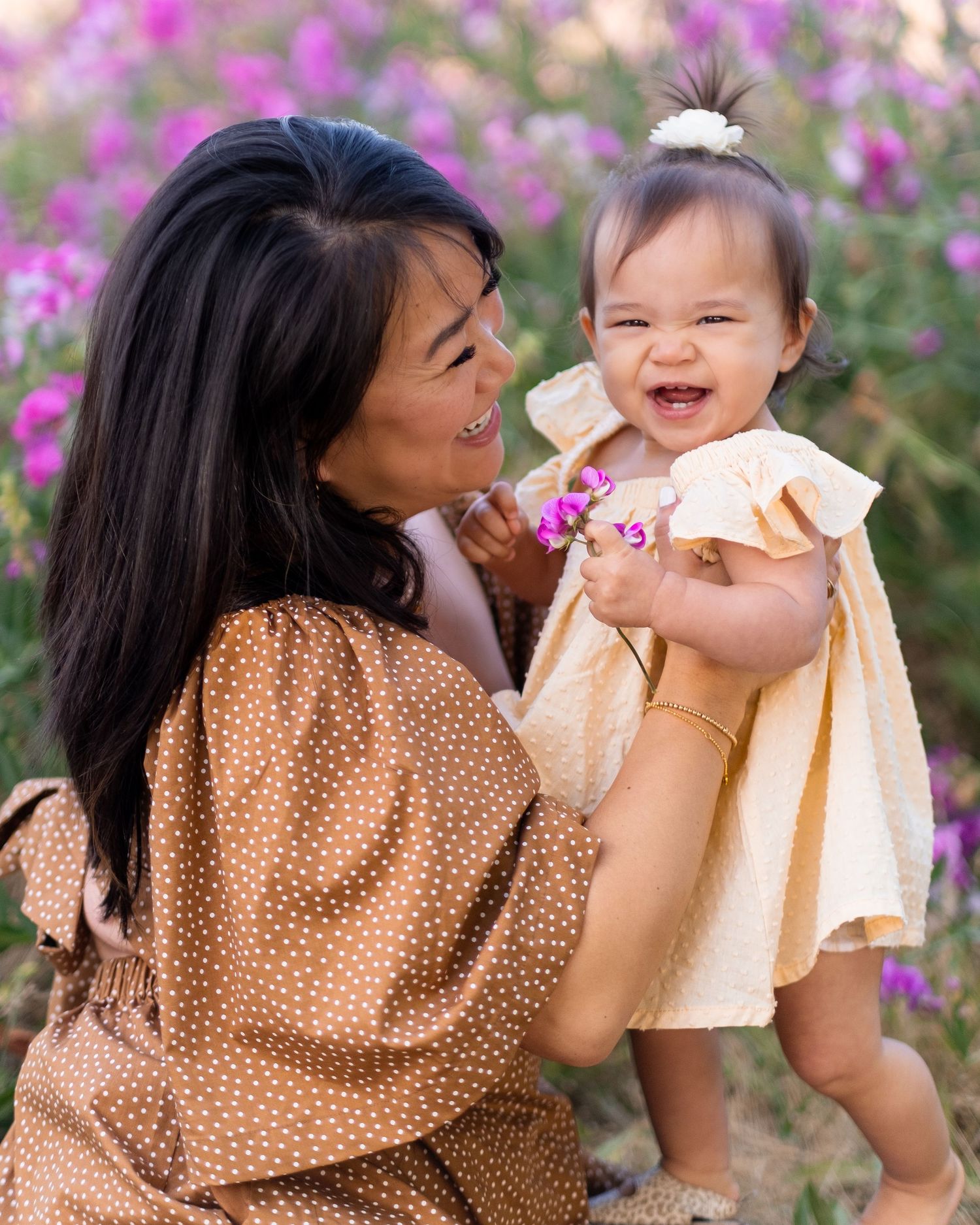 Two people in polka dot and beige outfits share a joyful moment against a backdrop of pink flowers in a garden.