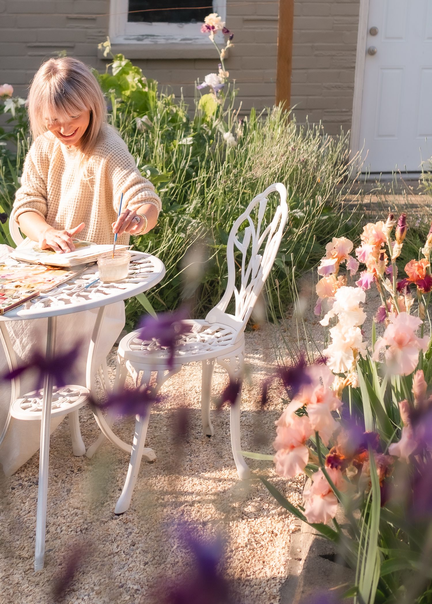 Woman painting at a white bistro table surrounded by blooming flowers in a sunny garden.