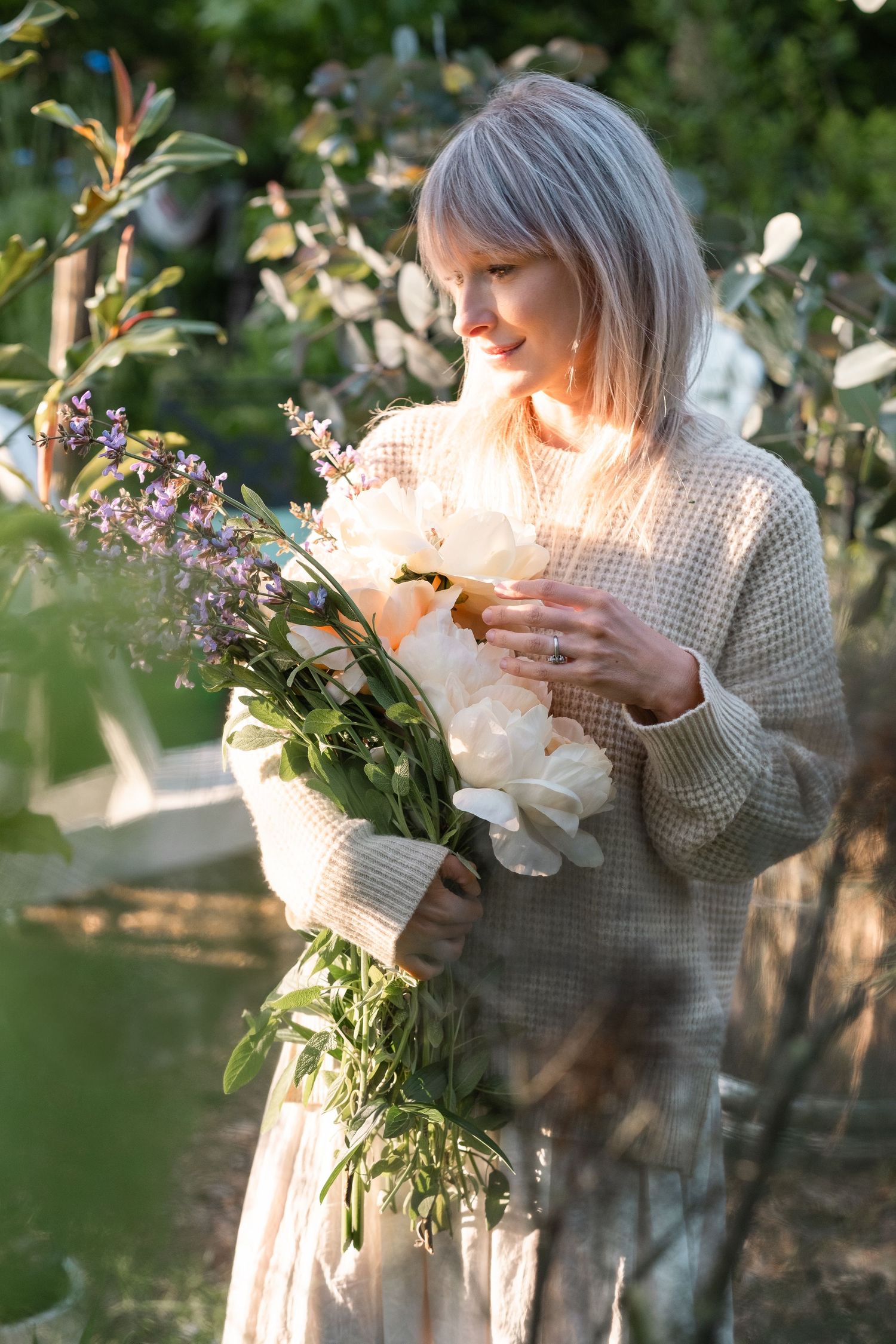 Woman in cream sweater holding white peony bouquet with lavender in golden sunlit garden.