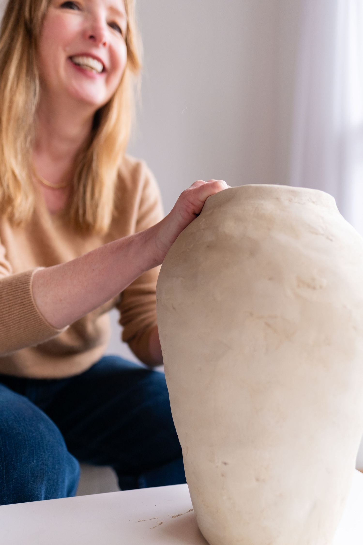 An artist working on a large white ceramic vase on a table in natural lighting.