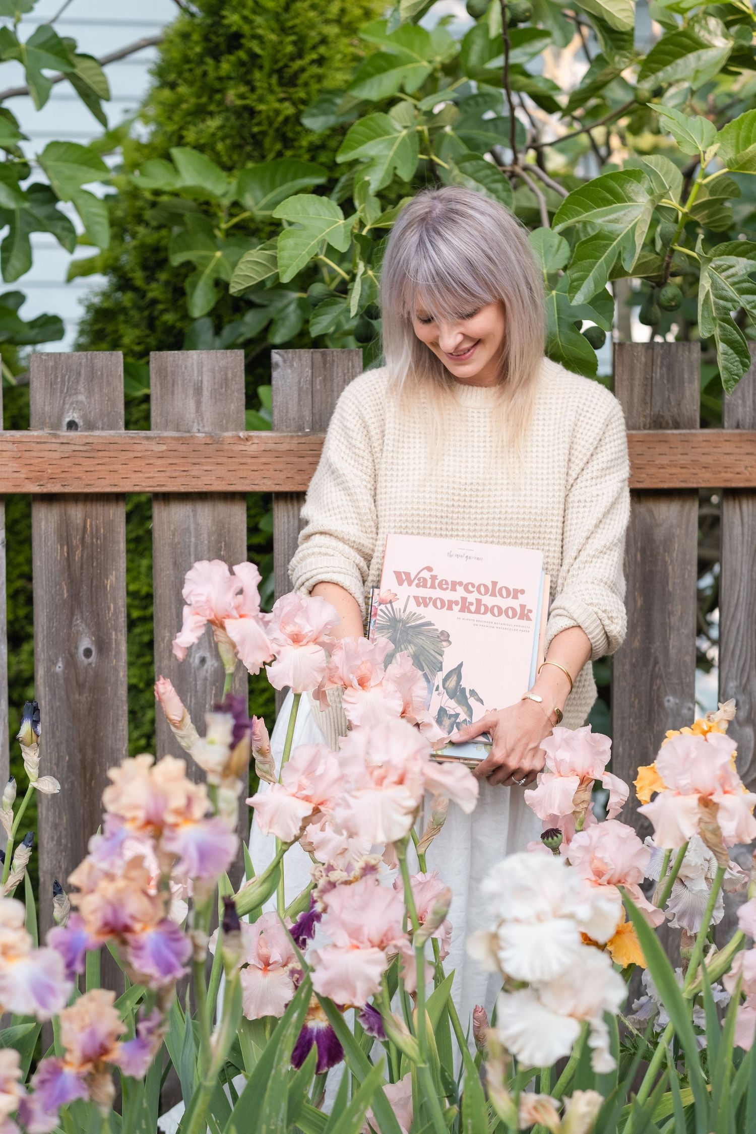 Smiling woman holding a Watercolor Workbook book among colorful iris flowers by a wooden fence.