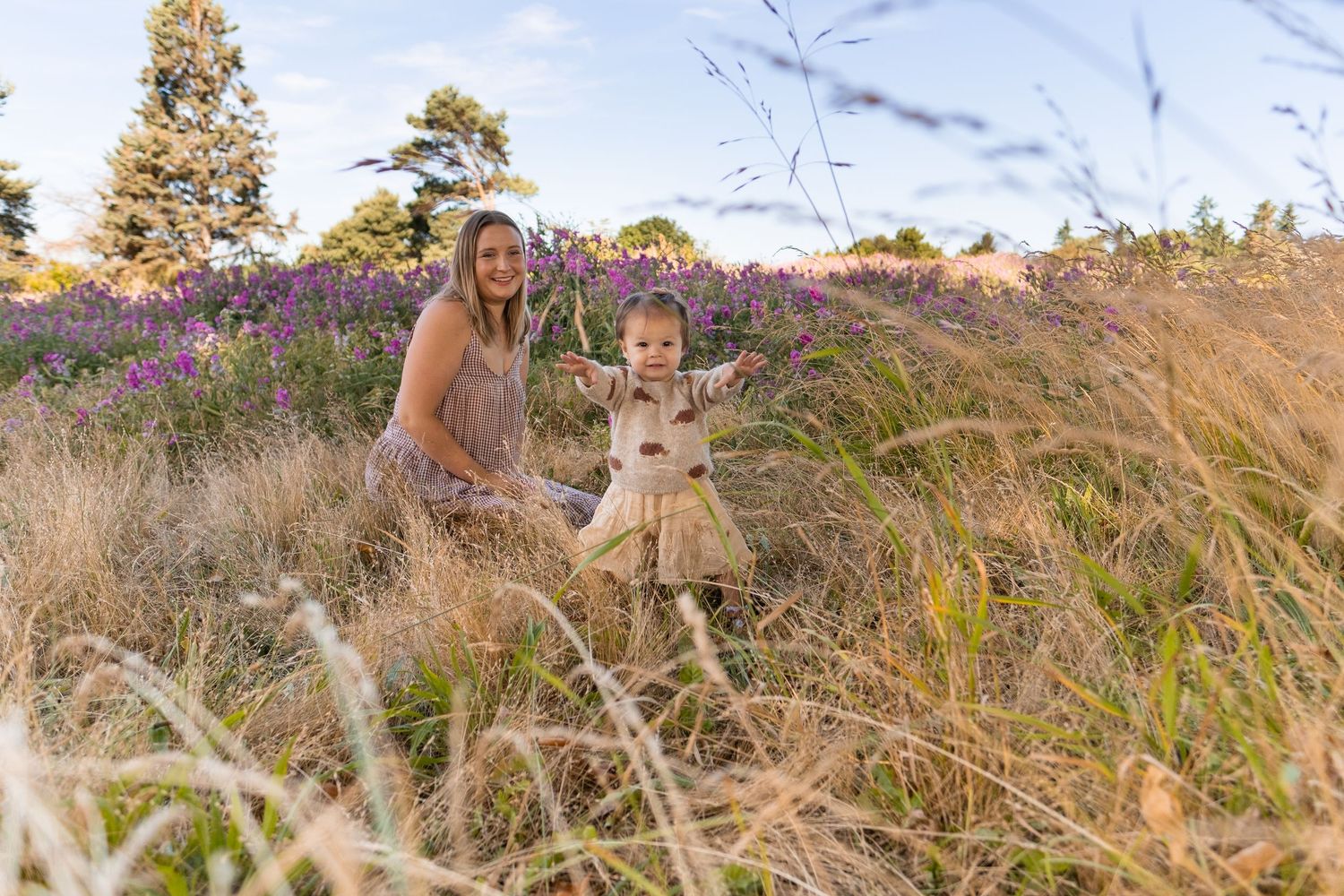 Two people stand together in a field of tall golden grass and purple wildflowers on a sunny afternoon.