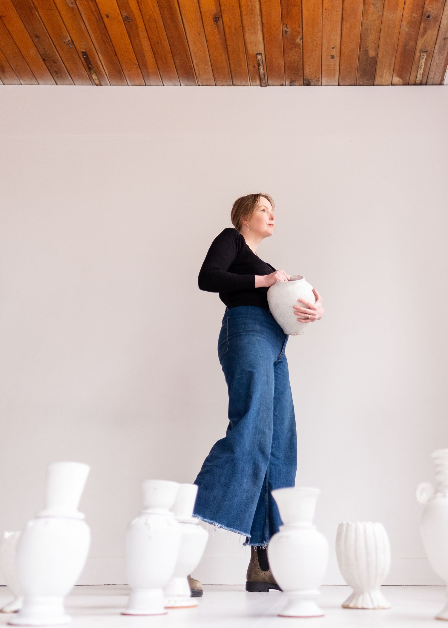 Someone in a black top and wide-leg jeans poses among white ceramic vases against a white wall with wooden ceiling.