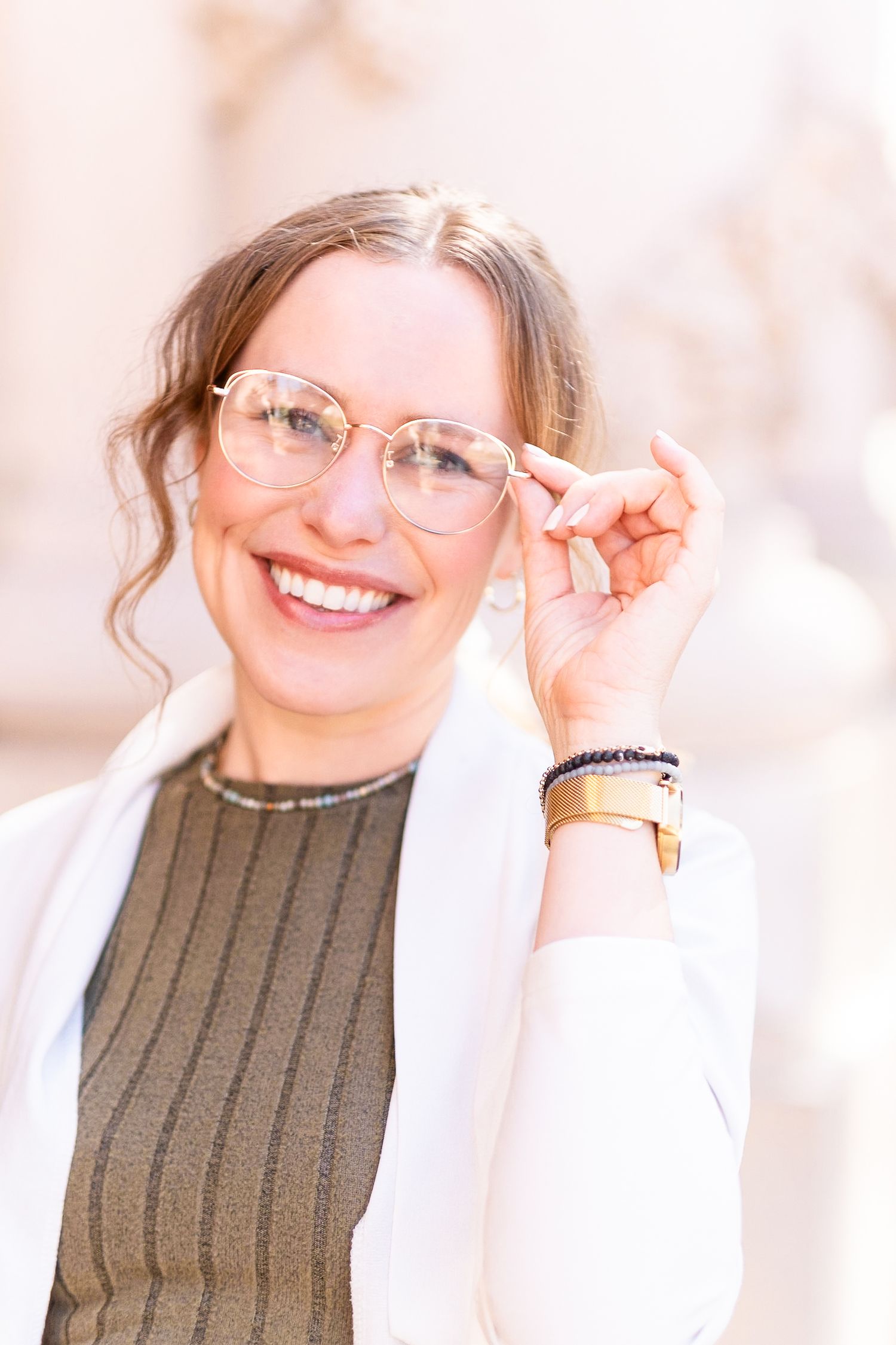 Smiling person wearing glasses and a brown sweater adjusts eyewear while wearing a white cardigan.