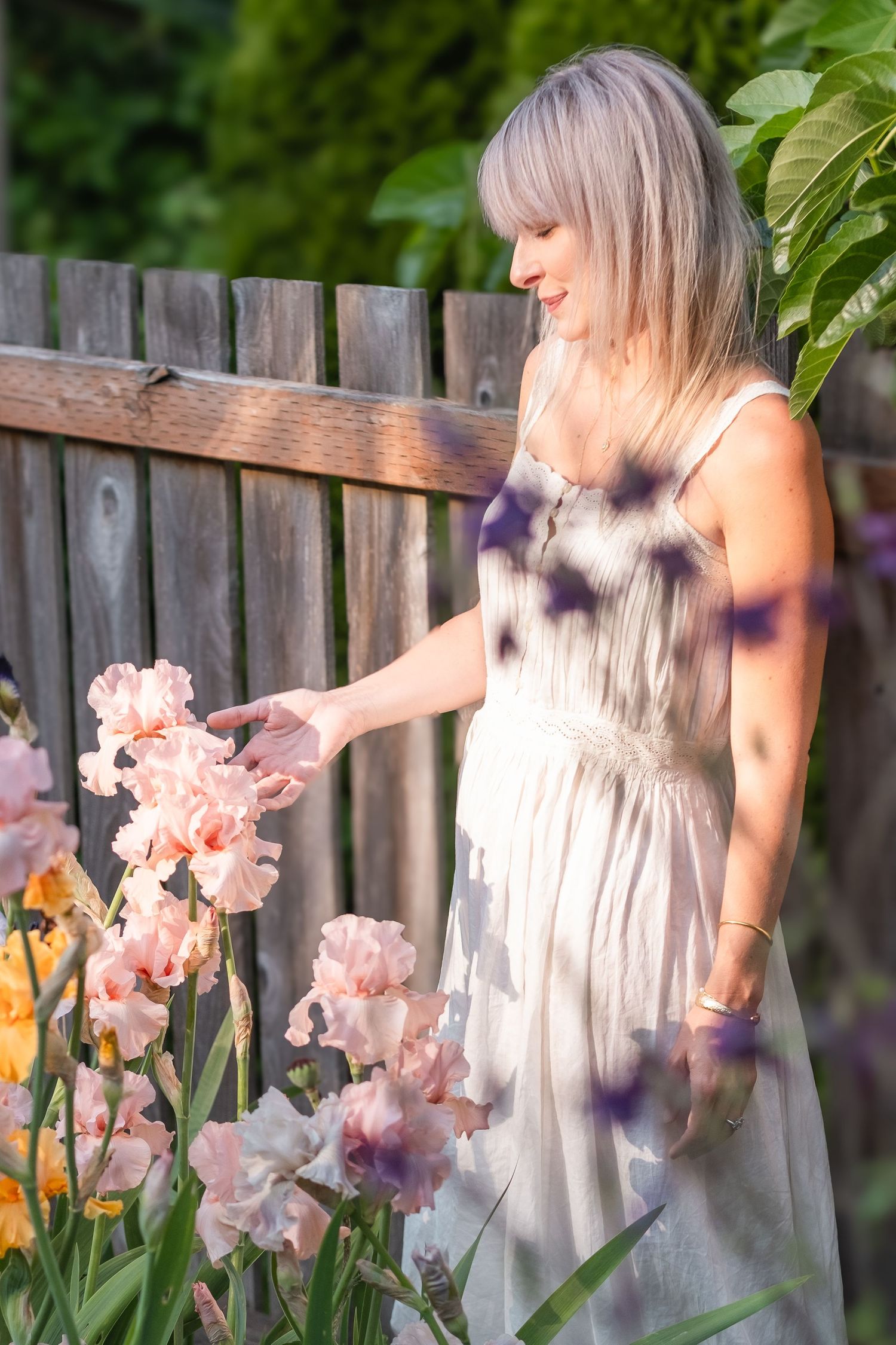 Blonde woman in white dress touching pink iris flowers by wooden fence.