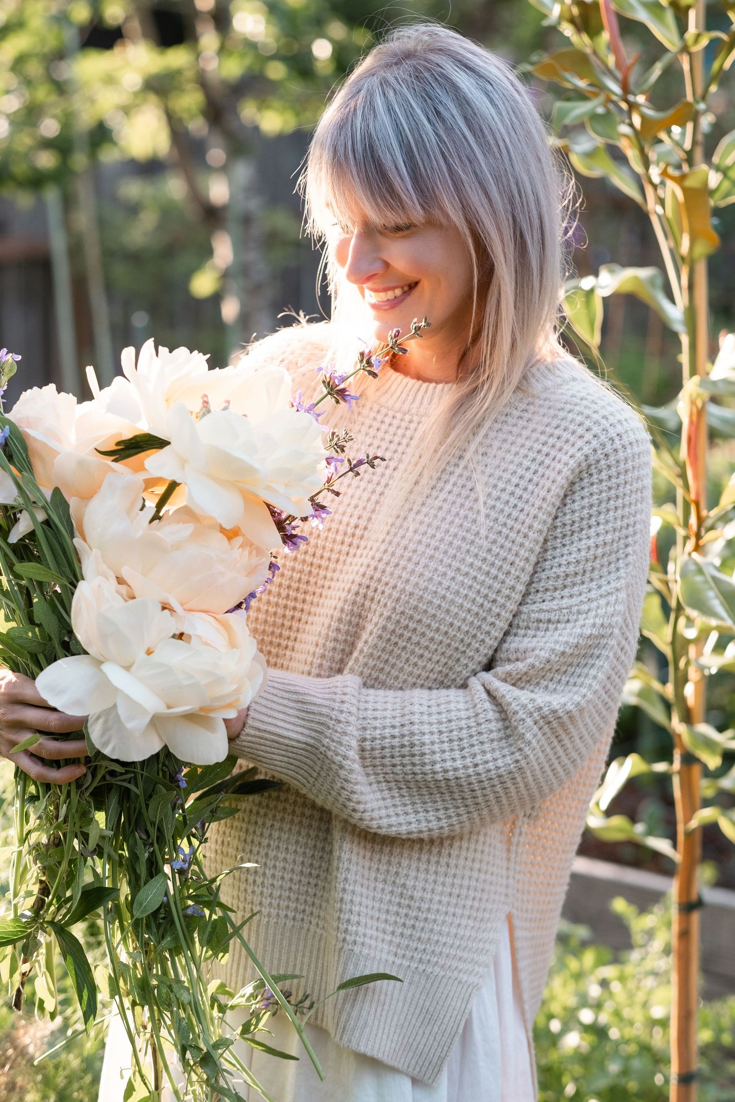 Smiling woman in waffle knit sweater holding large bouquet of white peonies in garden.