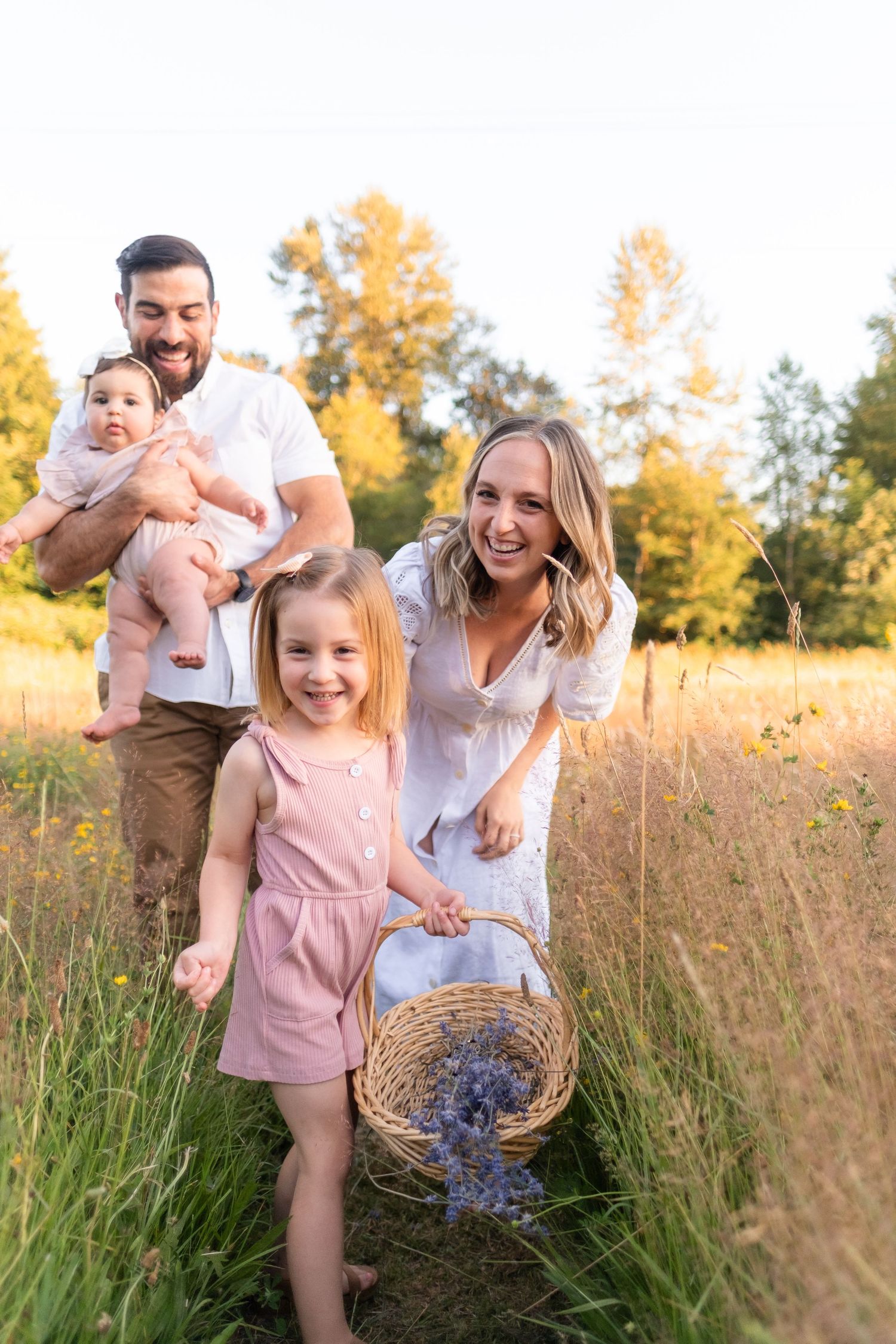 Happy family with baby and young girl walking through sunny meadow, carrying basket of lavender flowers.