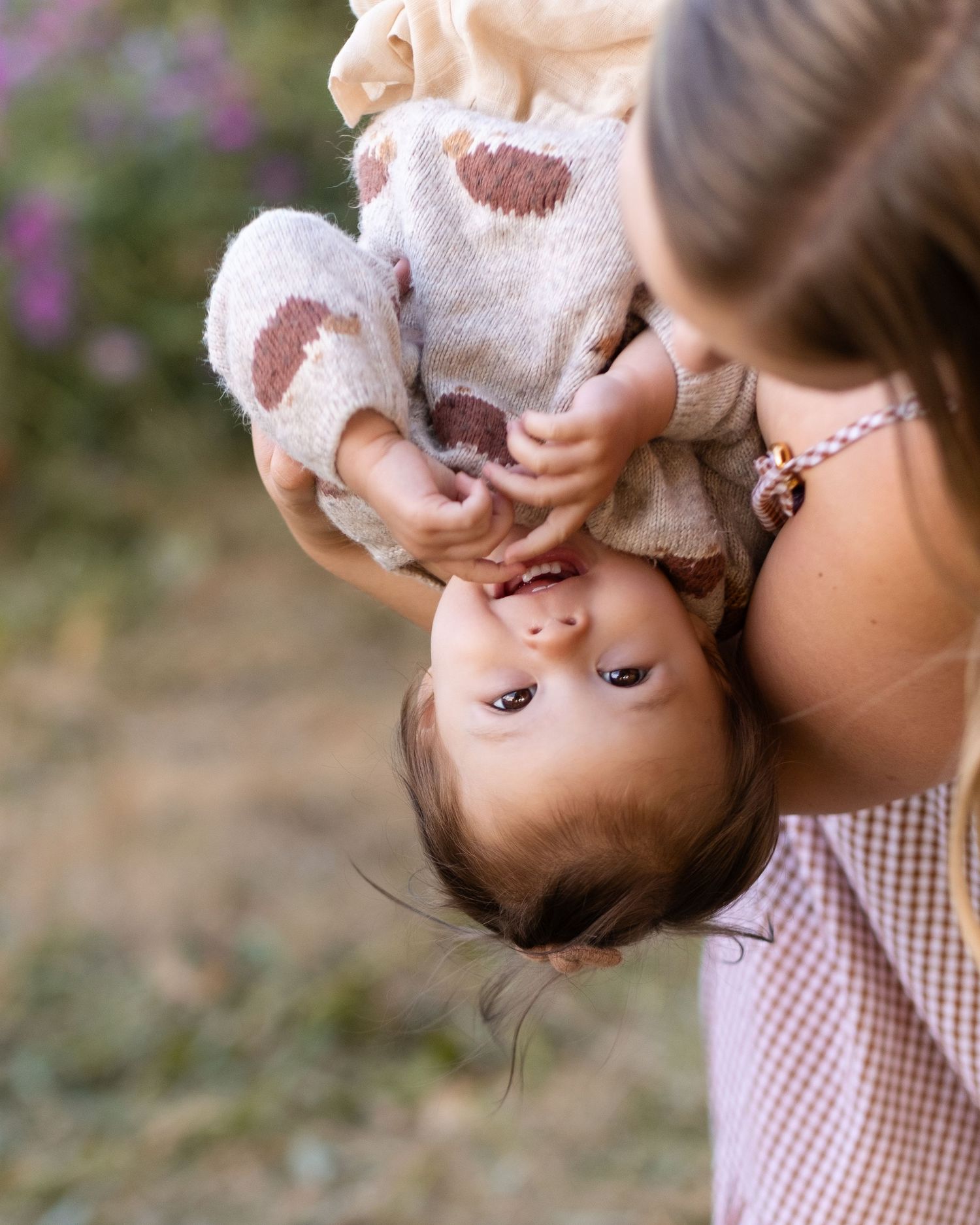 A young infant being held upside down in a playful moment wearing a white outfit with a gingham print dress visible.