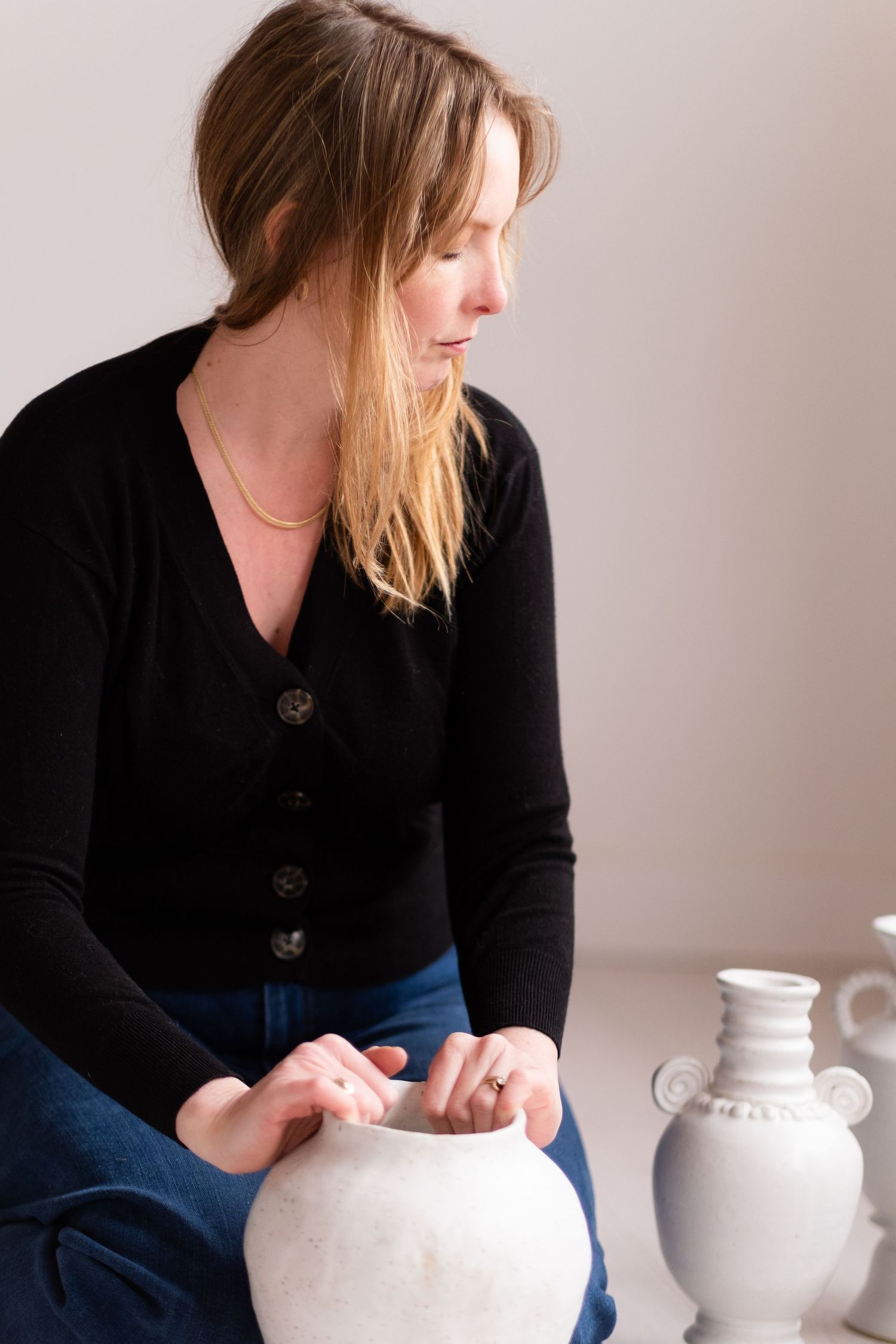 Person in dark sweater carefully handles white ceramic vessel against neutral backdrop with pottery collection.