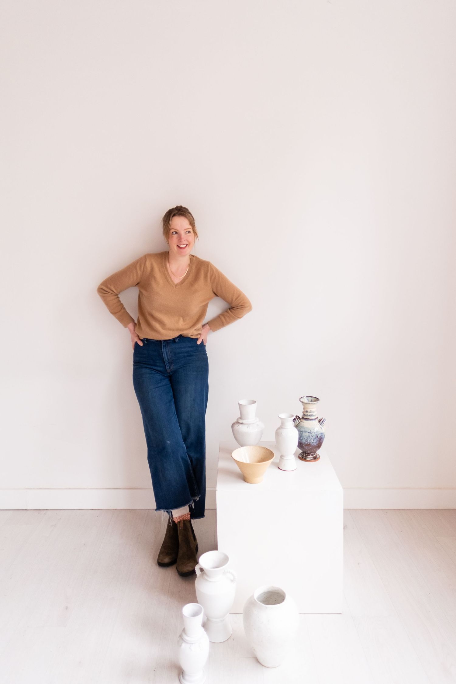Artist poses with collection of white ceramic vessels displayed on pedestal in bright minimal studio.
