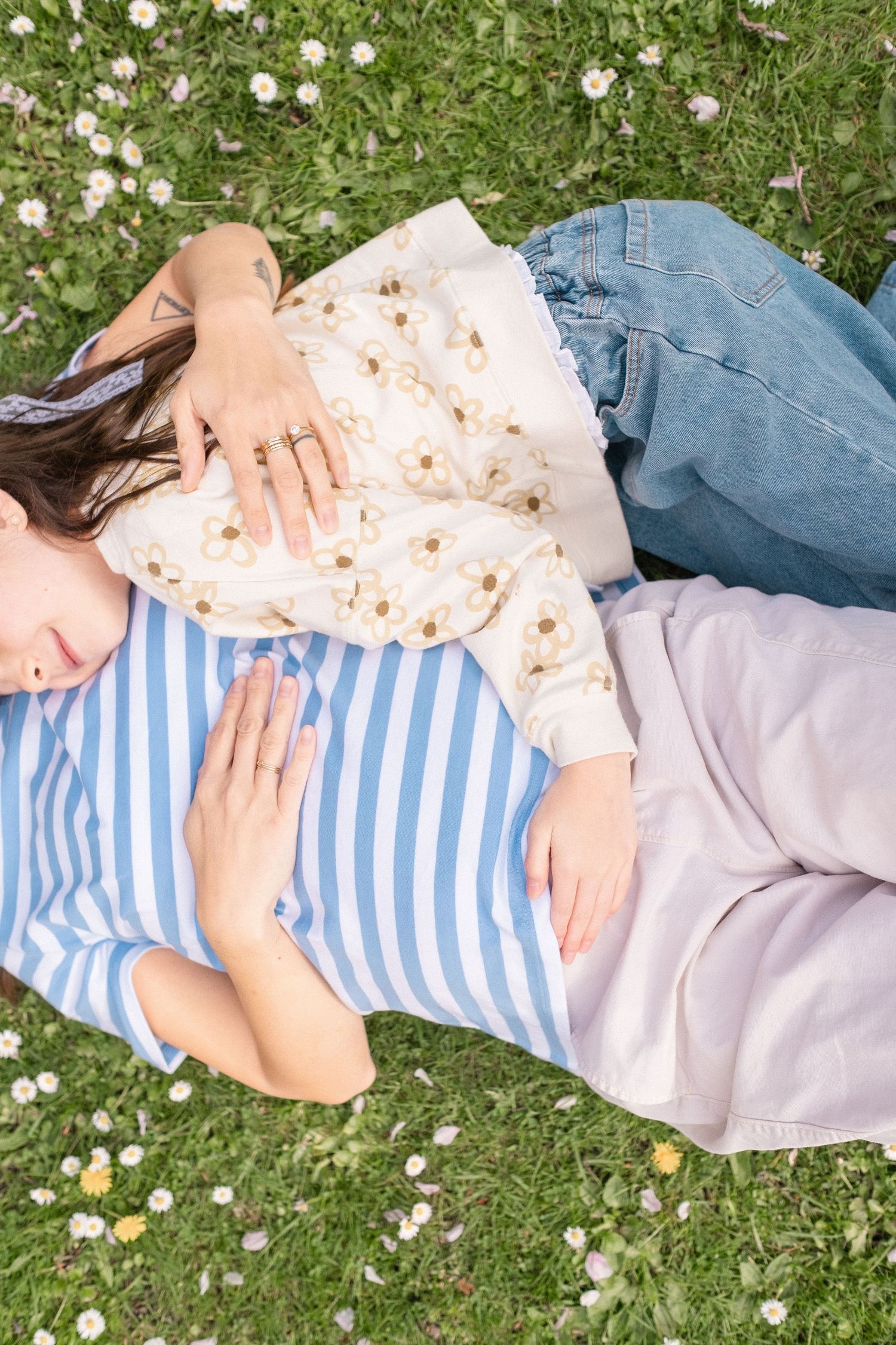 Two people lying on grass surrounded by daisies during springtime, wearing casual clothes.