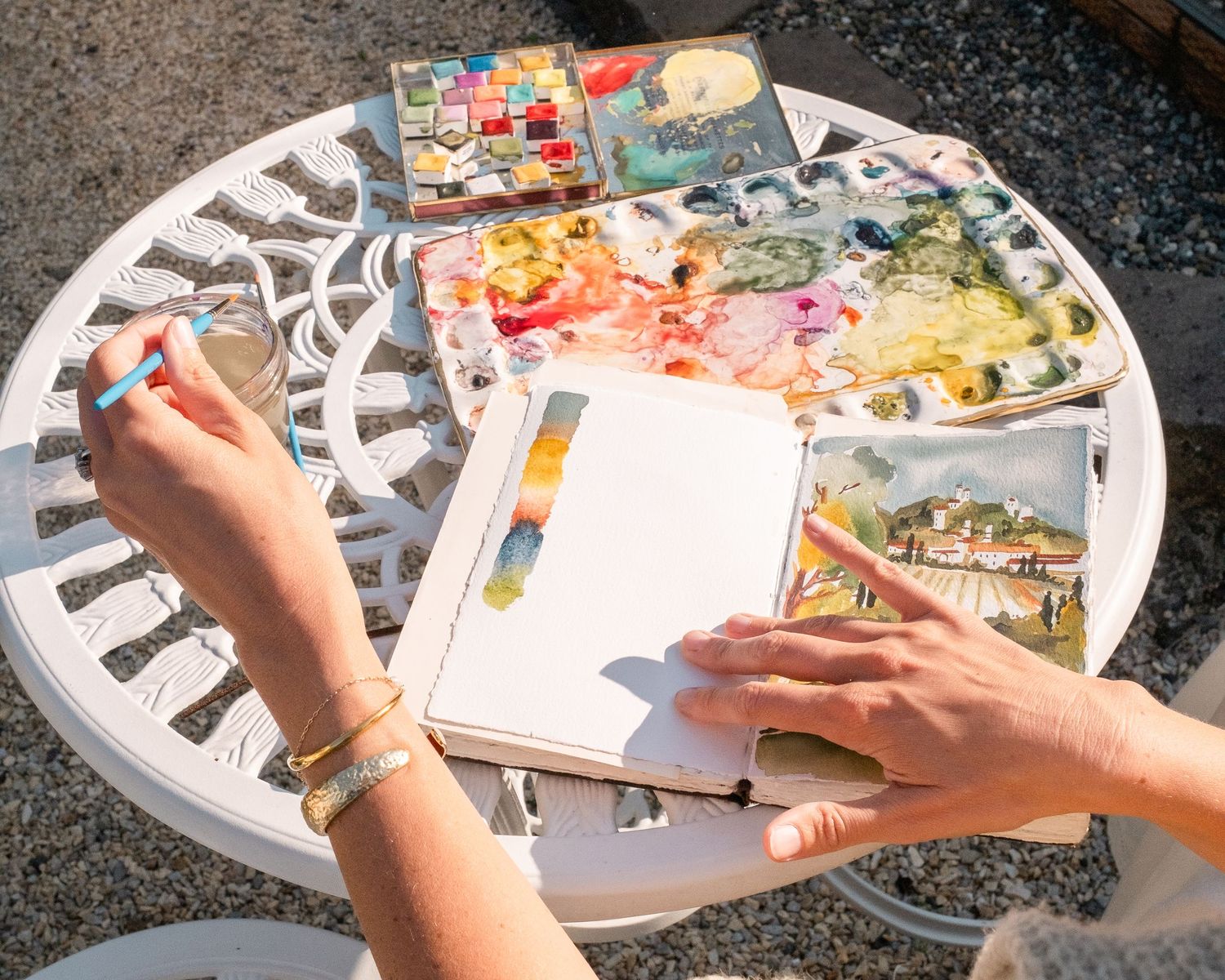 Artist painting watercolors at a white outdoor table with paint palette, brushes, and sketchbook in natural sunlight.