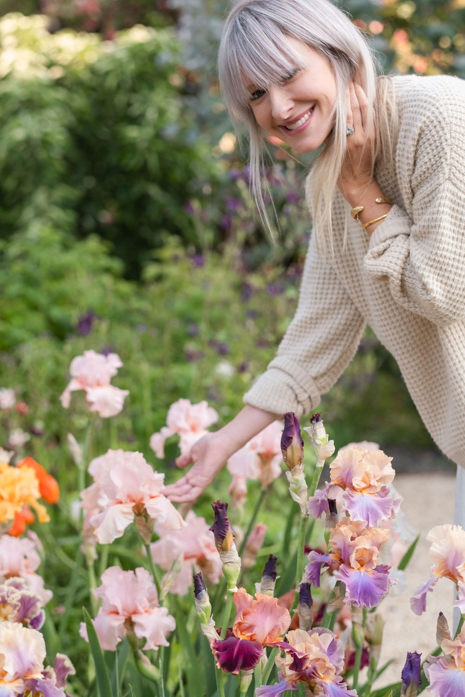 Smiling woman in cream knit sweater tending to pink and purple iris flowers in lush garden.