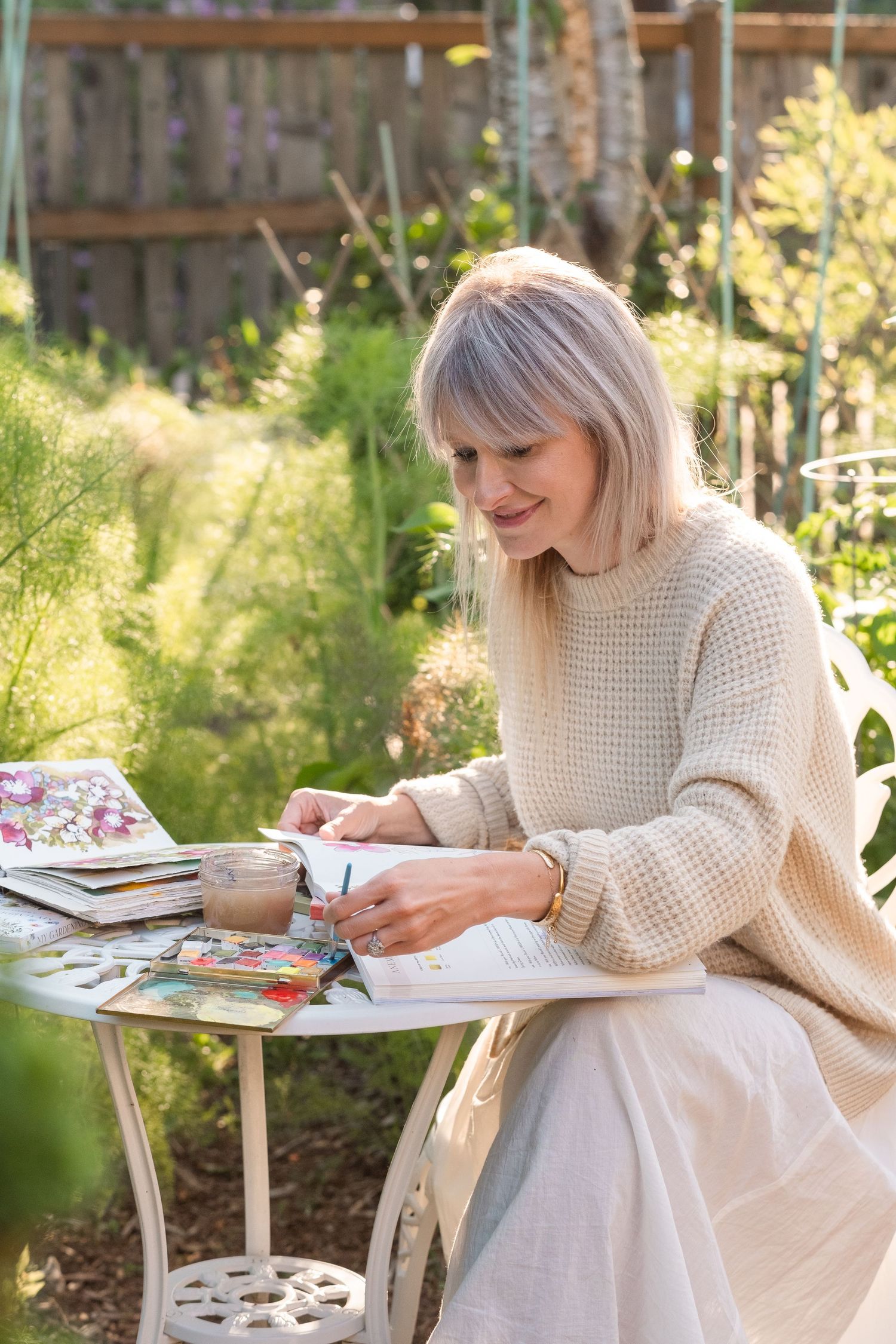 Smiling blonde woman in cream knit sweater writing in a gardening journal at a white garden table.