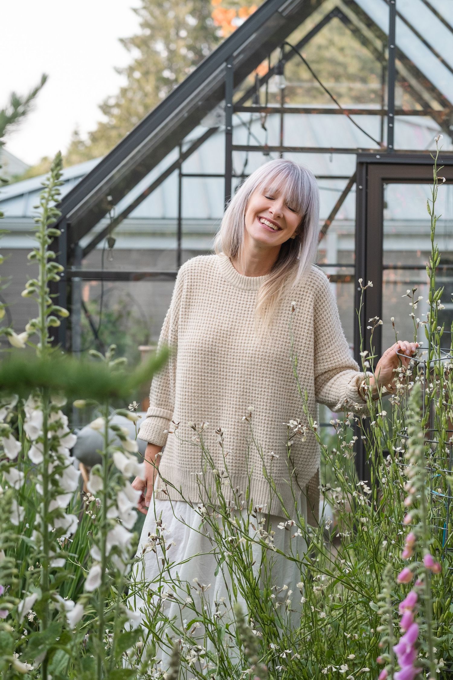 Cheerful woman in beige sweater laughing among wildflowers with a glass greenhouse in the background.