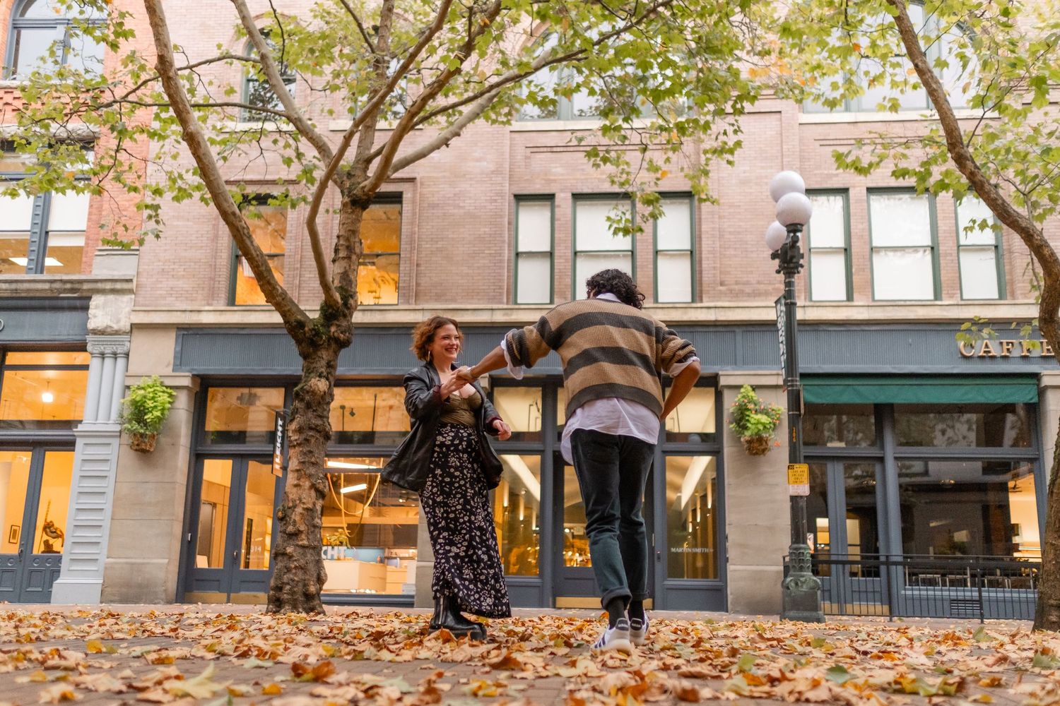 Couple dancing joyfully on a leaf-covered sidewalk in autumn, surrounded by charming brick storefronts.