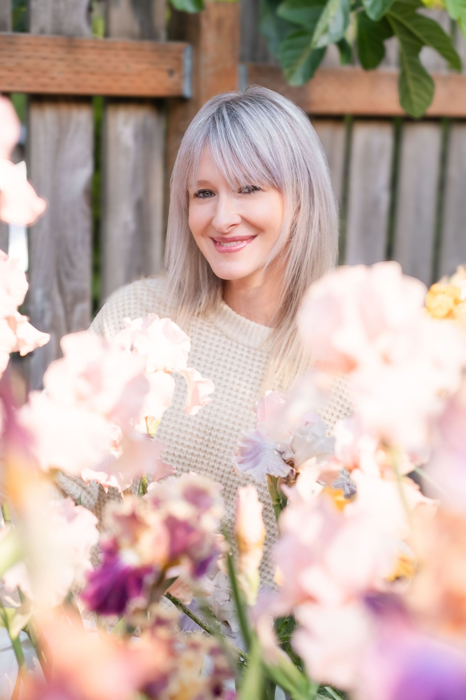 Smiling blonde woman surrounded by soft pink and purple iris flowers in a sunny garden.
