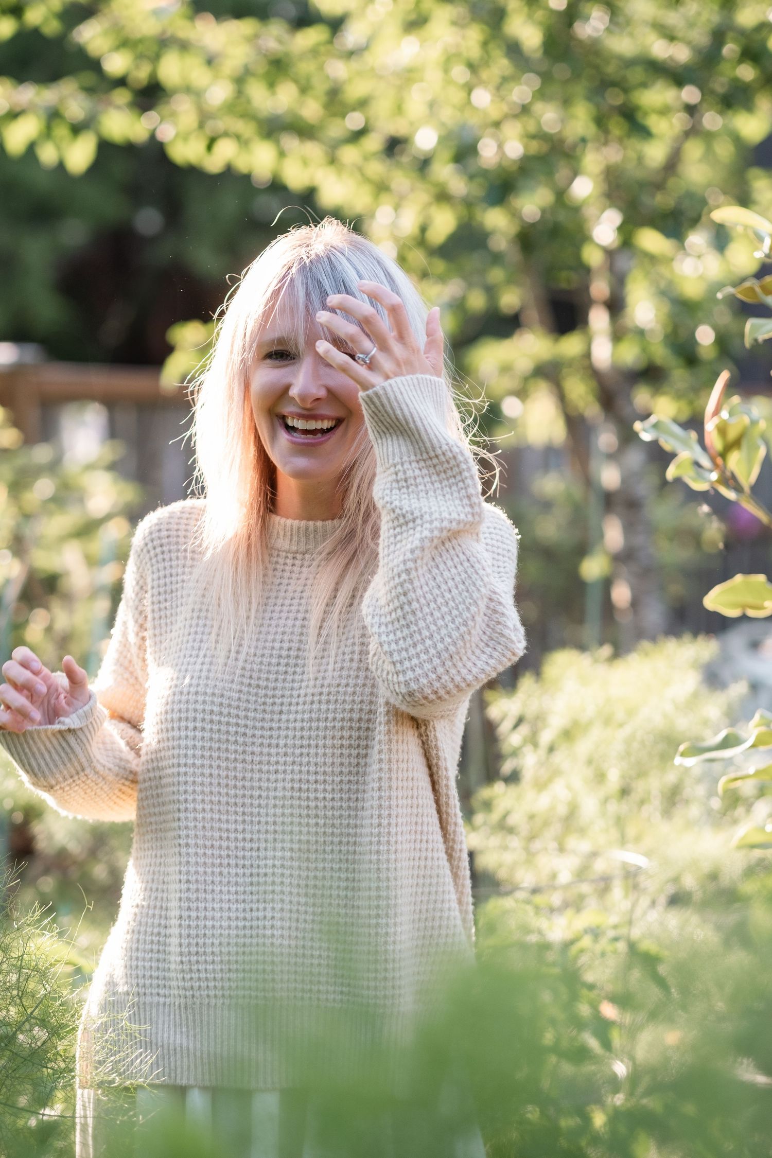 Happy older woman with gray hair laughing outdoors in a garden, wearing a cream waffle knit sweater.