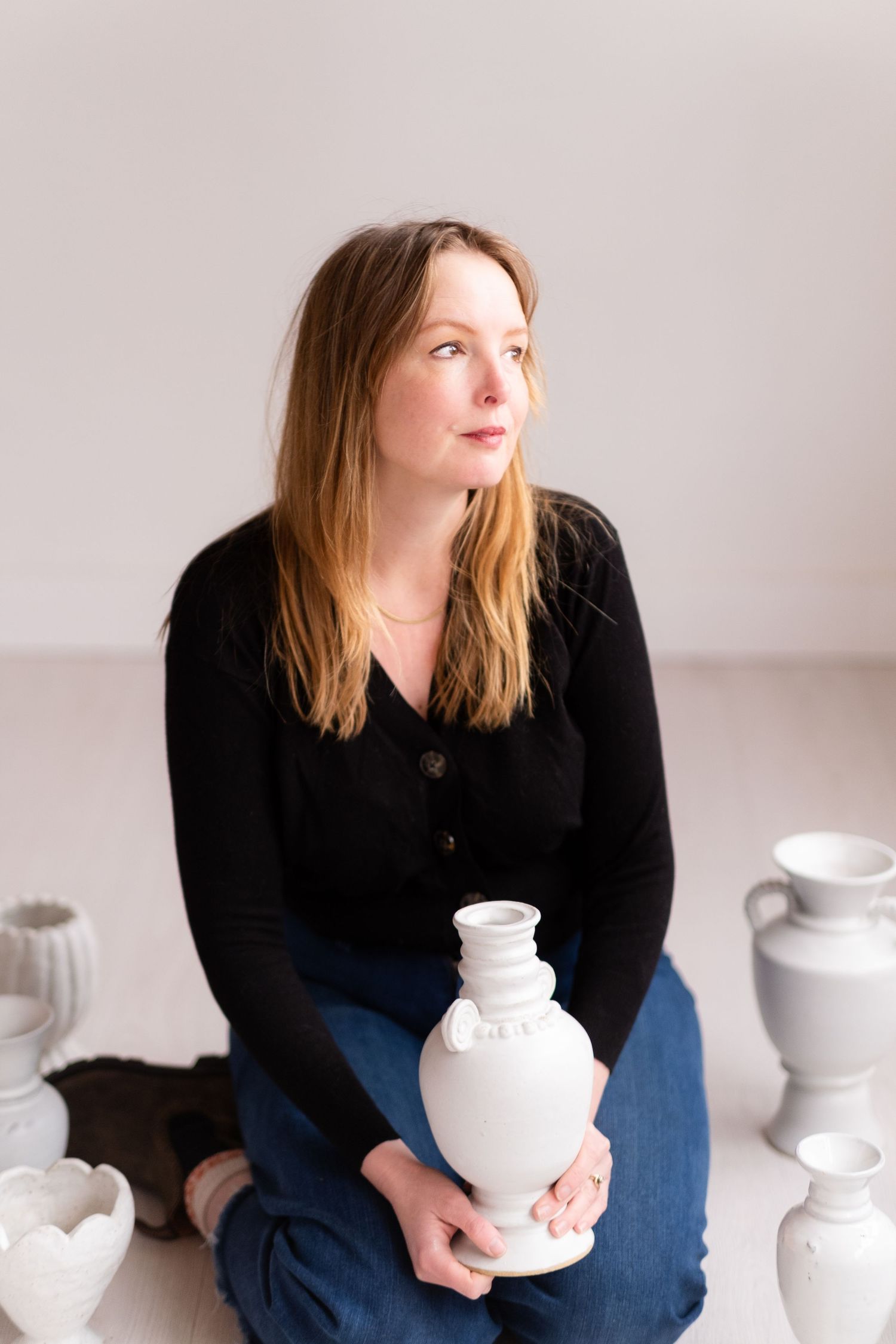 Ceramic artist in black sweater holds white vase while sitting among pottery collection.