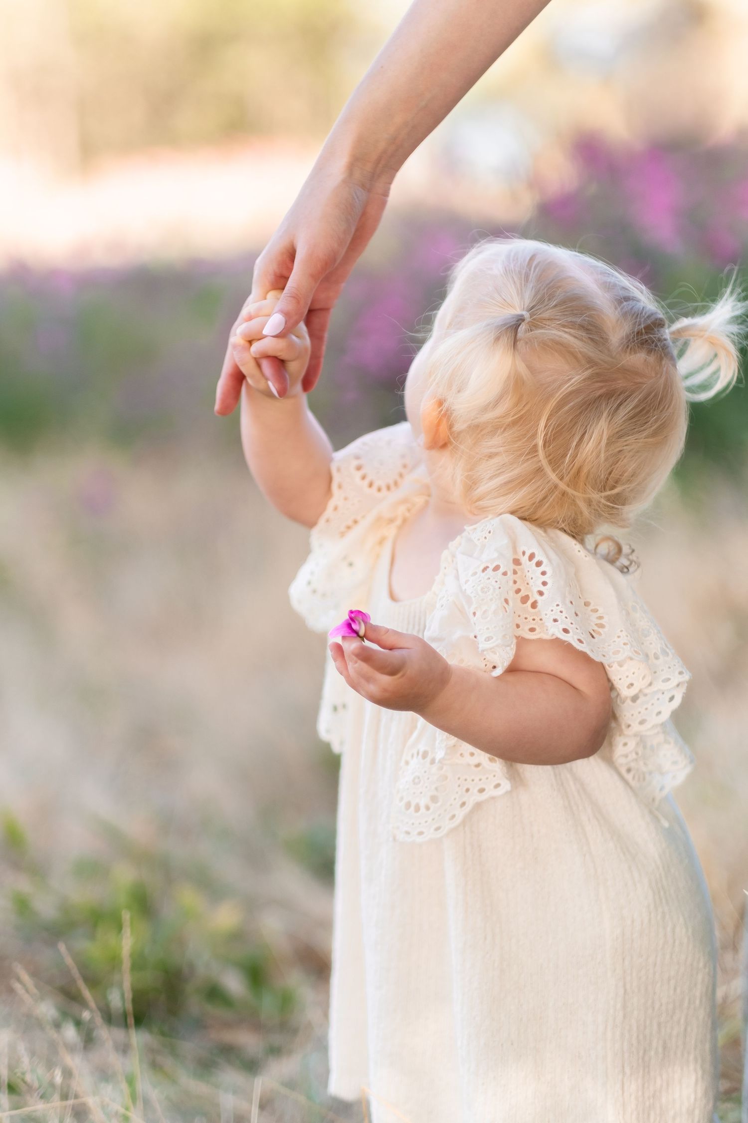 A small child in a white eyelet dress holds hands with someone while standing among pink flowering plants.