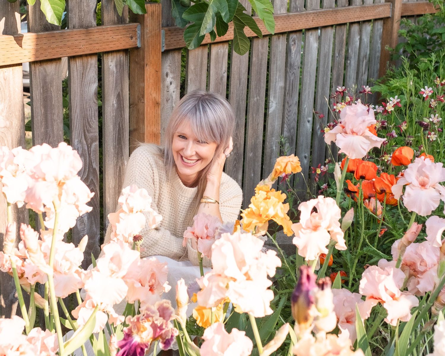 Woman with silver hair smiling among colorful pink, orange, and red iris flowers in a wooden fence garden.