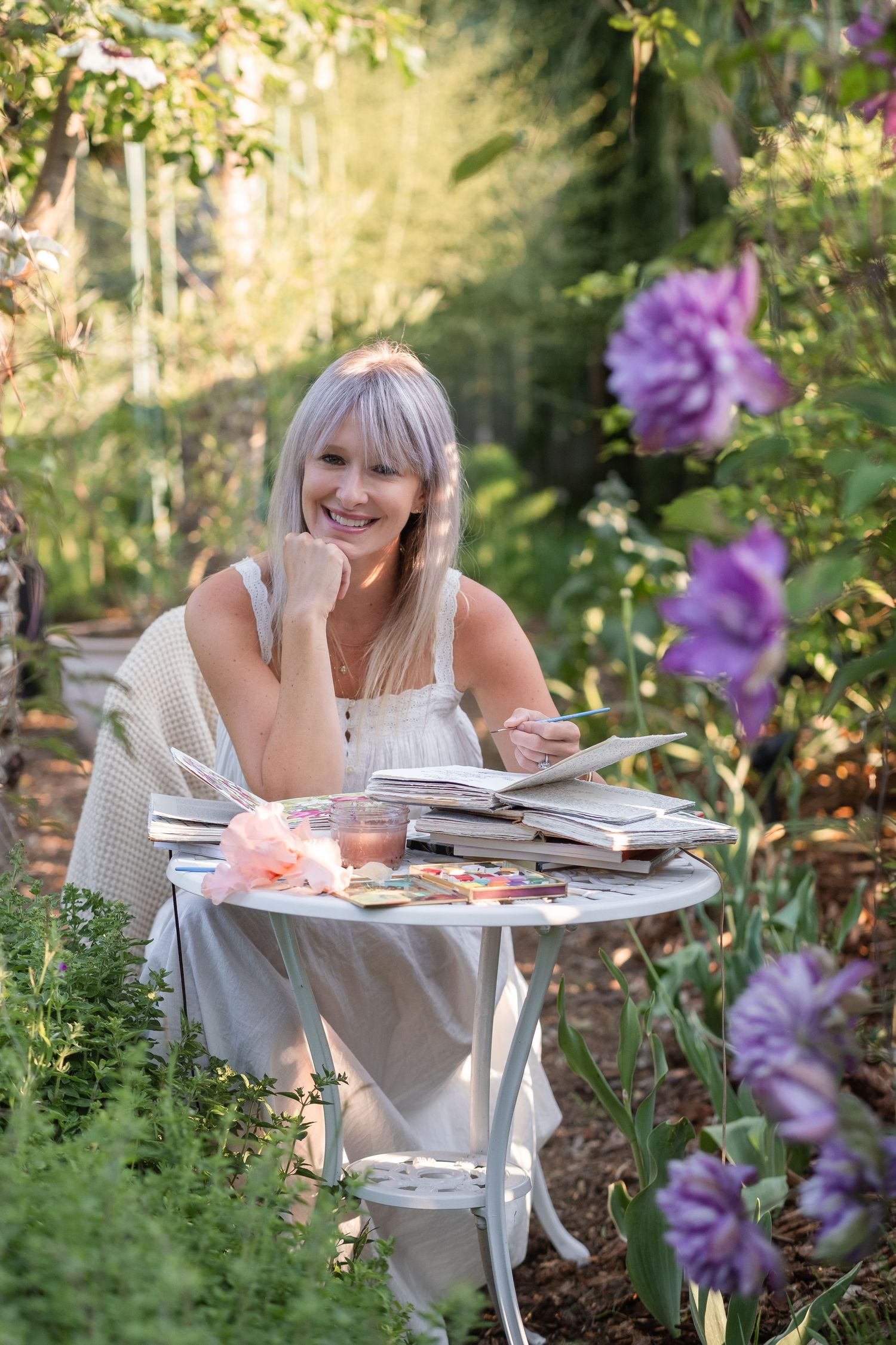 Smiling woman sitting at white garden table with books and art supplies among purple flowers.