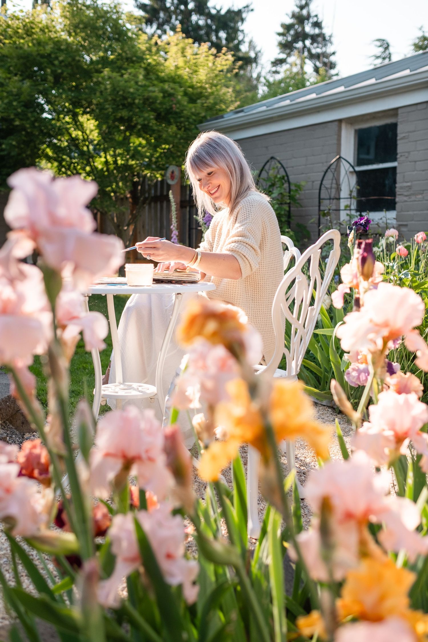 Smiling woman enjoying tea at a white garden table surrounded by blooming pink iris flowers.
