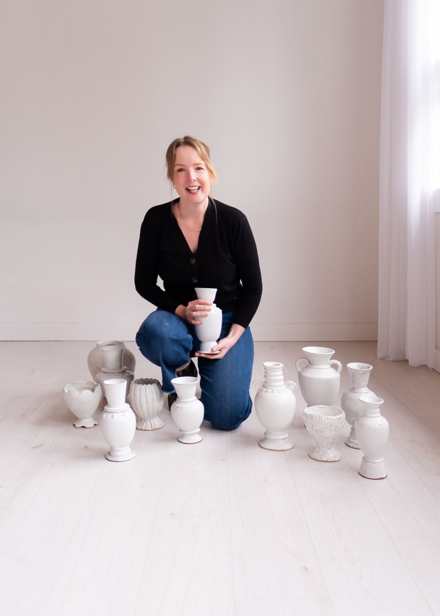 Ceramic white vases arranged on light floor with person in black sweater sitting among pottery collection.