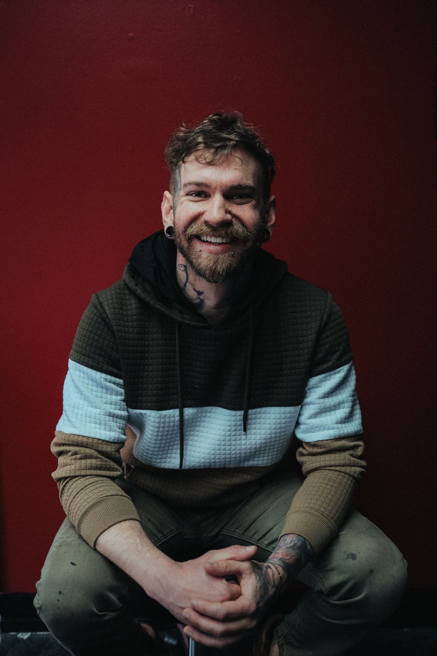 Person wearing a color-block hoodie sweater sits casually against a dark red background while smiling warmly.