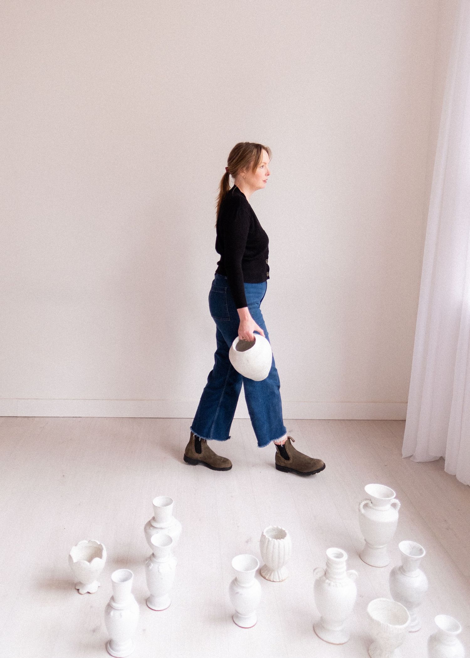 Person in dark clothing walking with white ceramic vases arranged on the floor.
