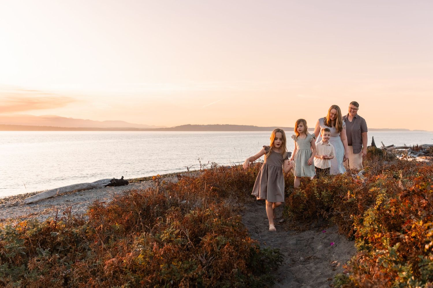 Family of five walking along a scenic beach at golden sunset with calm ocean waters and autumn foliage.