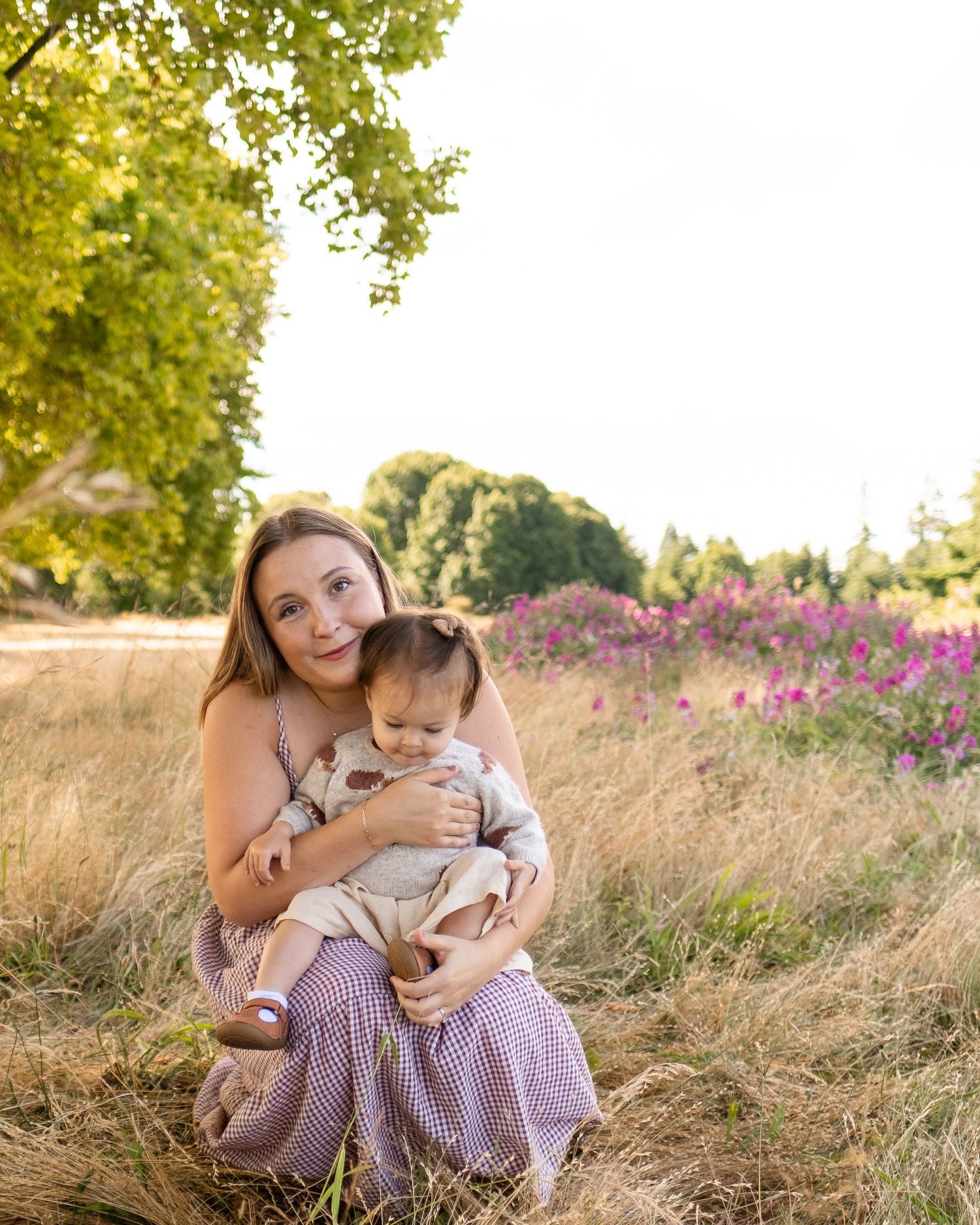 Parent and infant sit together in golden grass field with purple wildflowers and trees in summer afternoon light.