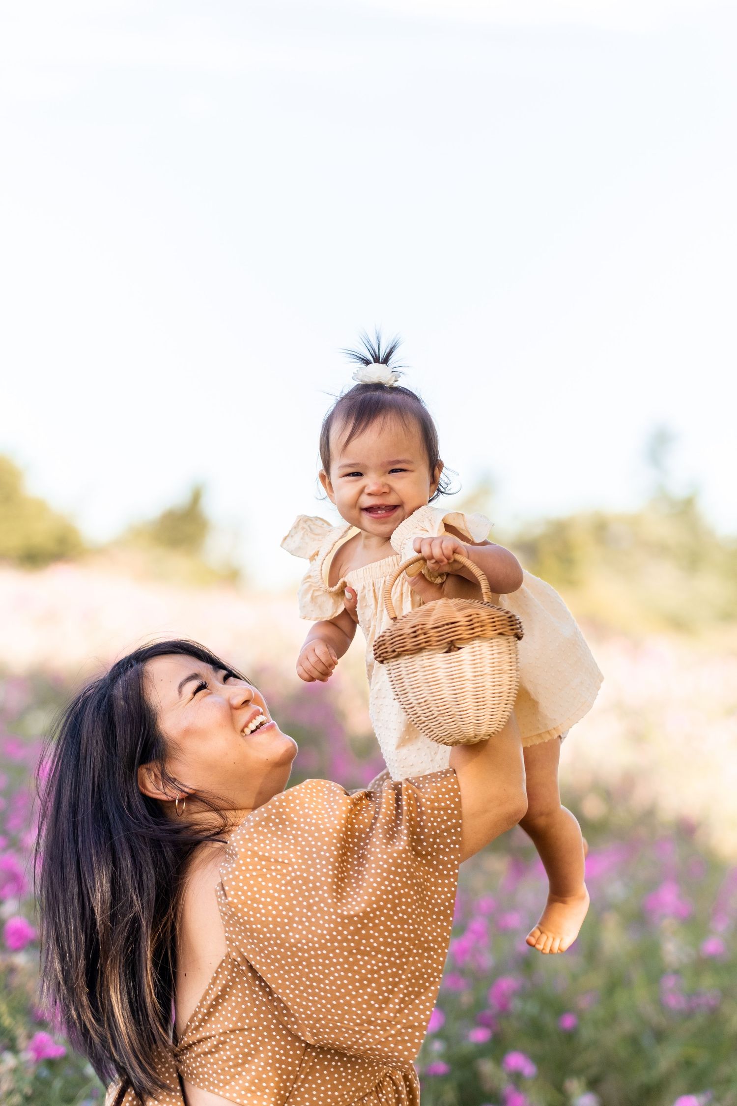 A parent lifts baby in a floral field during sunset while wearing a polka dot dress and sharing a joyful moment.
