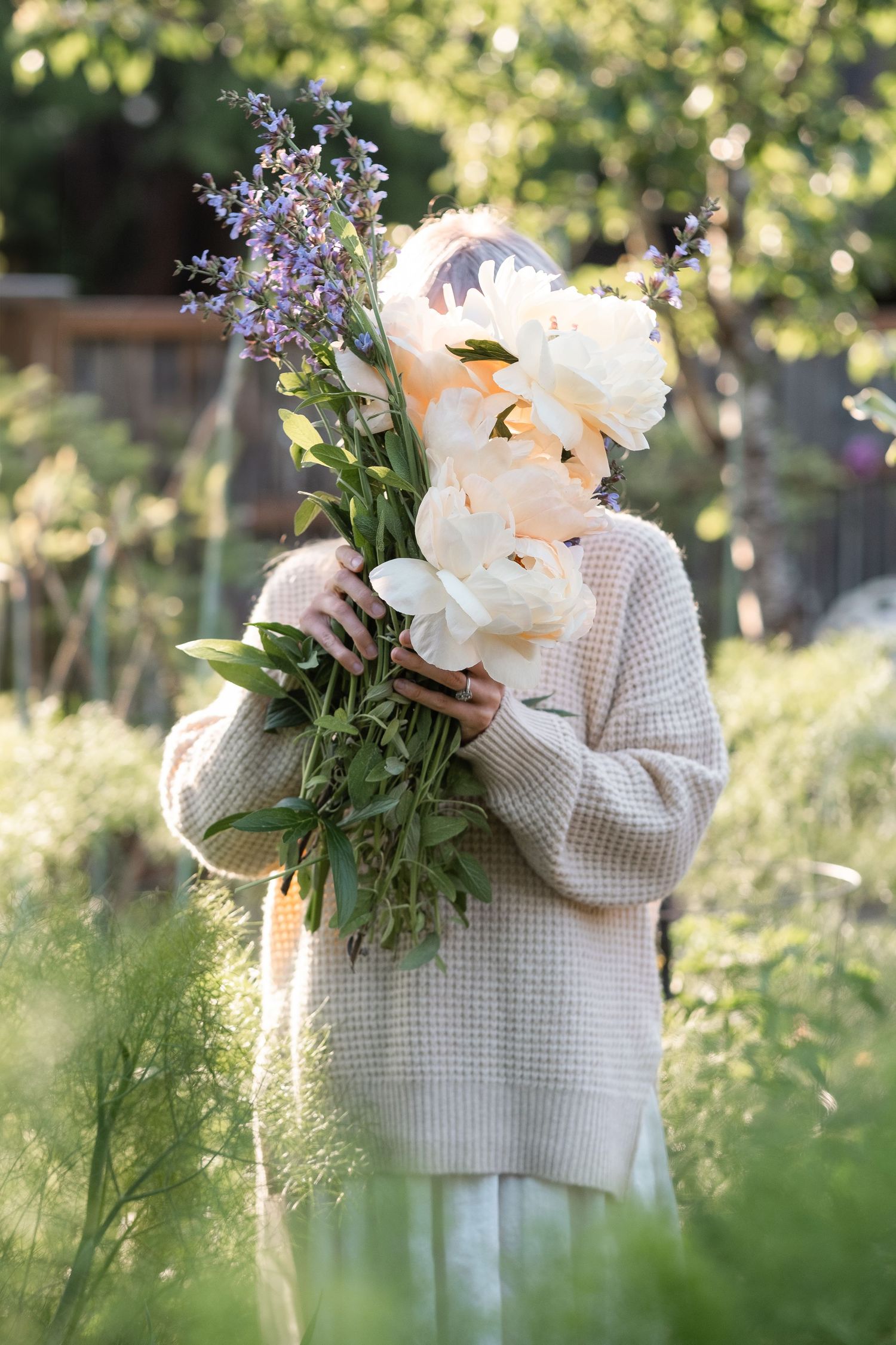 Person in beige sweater holding large bouquet of white peonies and lavender hiding their face.