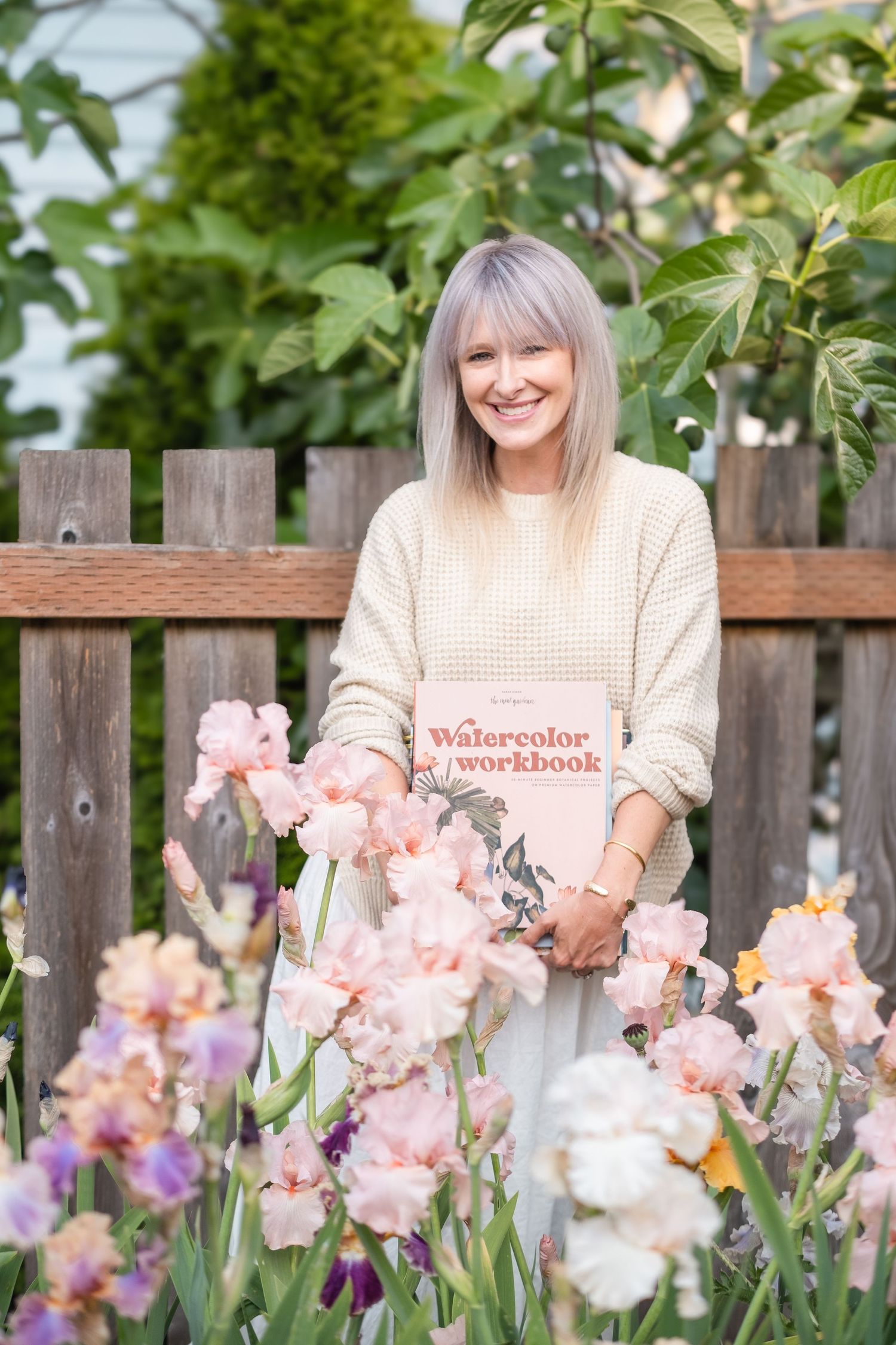 Smiling woman holding Watercolor Workbook book surrounded by blooming pink iris flowers outdoors.