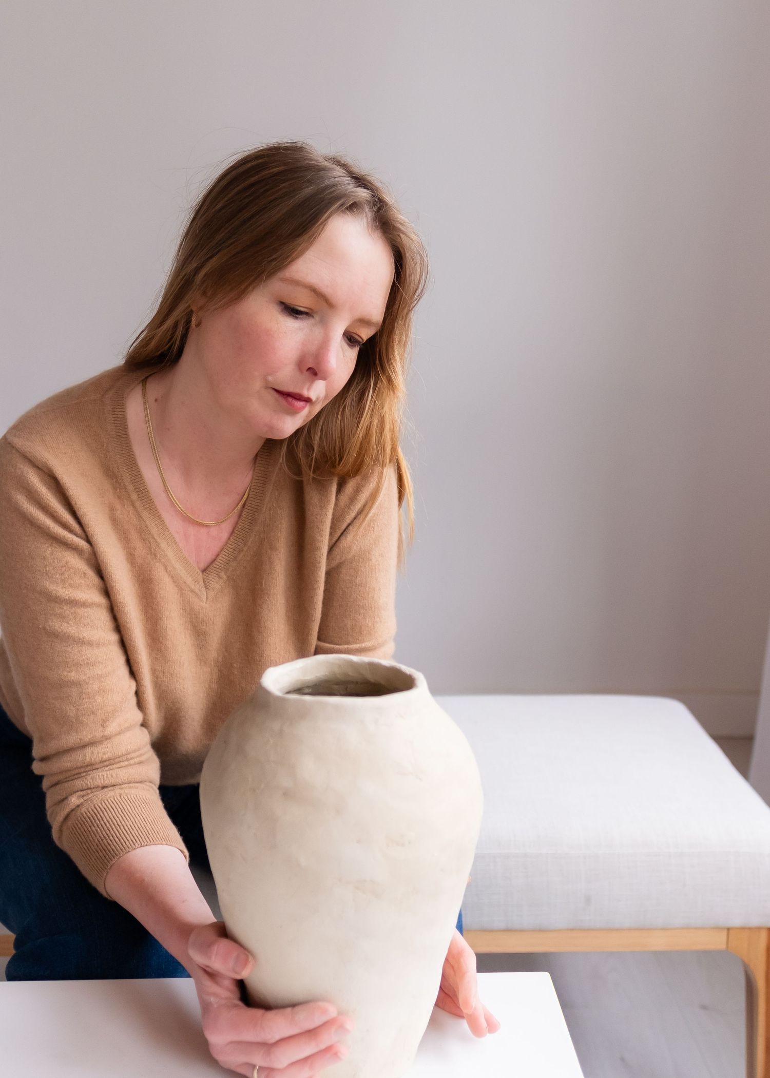 Person in tan sweater examines large cream pottery vessel on white table in minimalist setting.