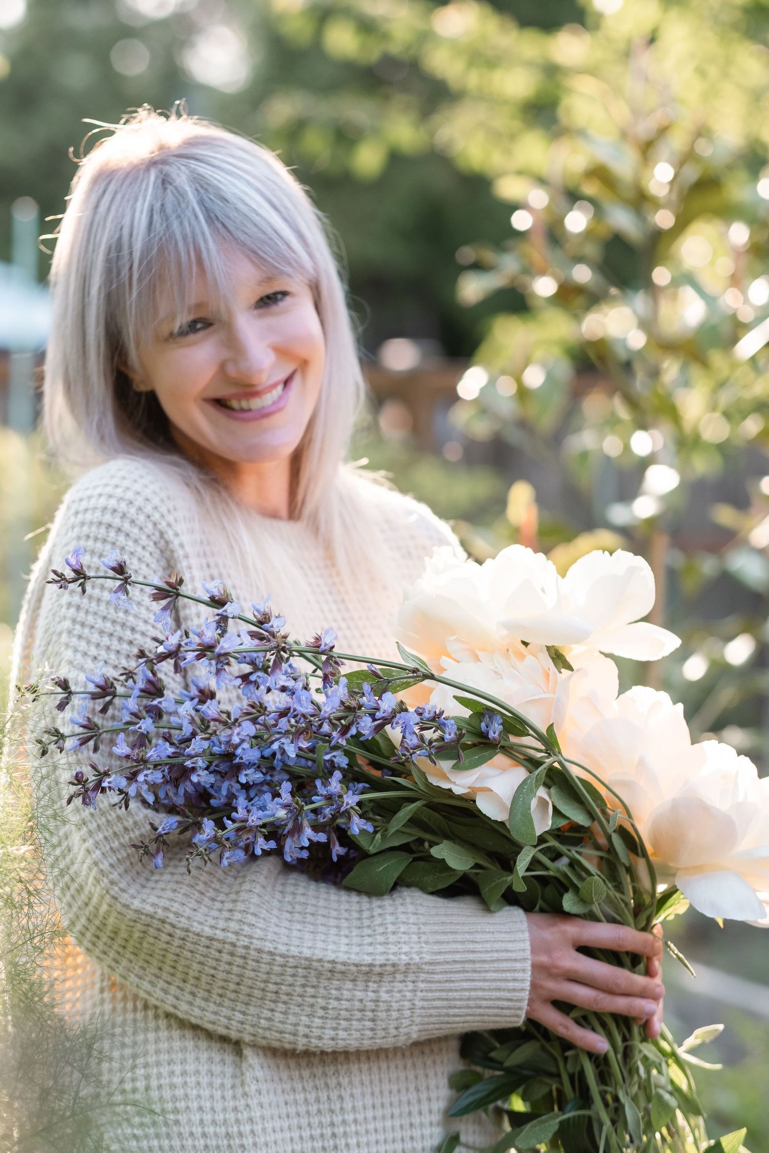 Smiling blonde woman in a cream sweater holding white peonies and purple wildflowers outdoors in a garden.