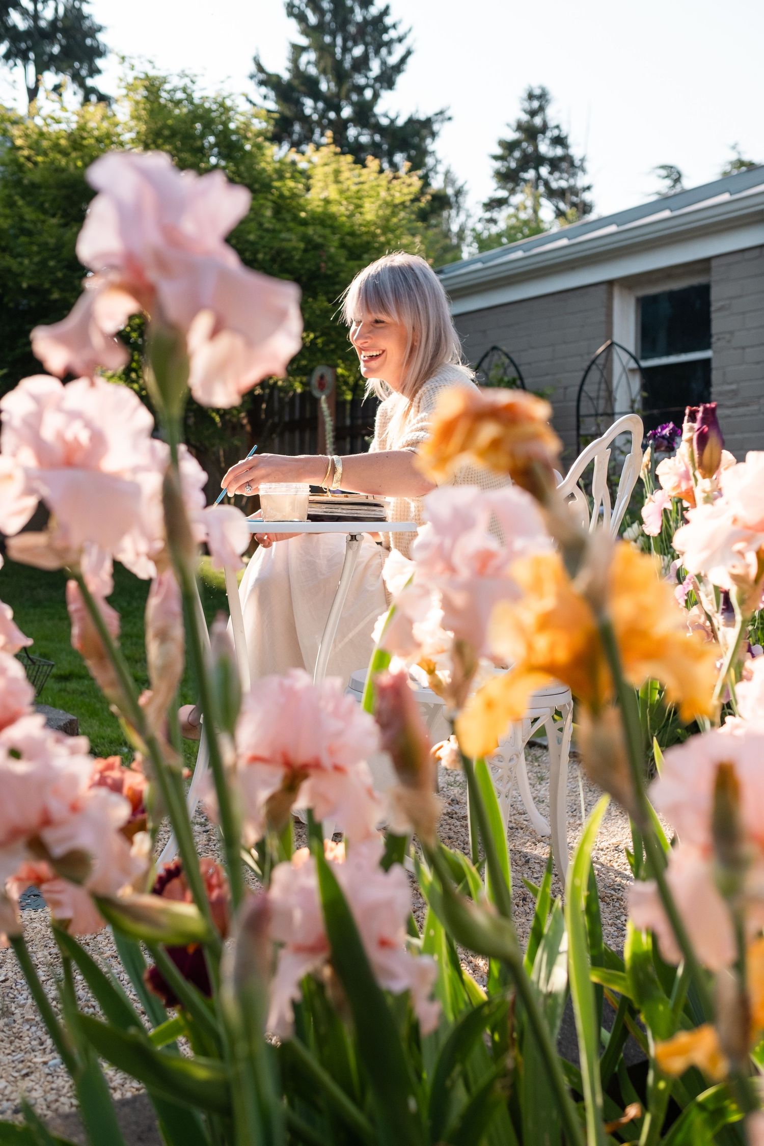 Blonde woman in white dress smiling among blooming pink and yellow iris flowers in a sunny garden.