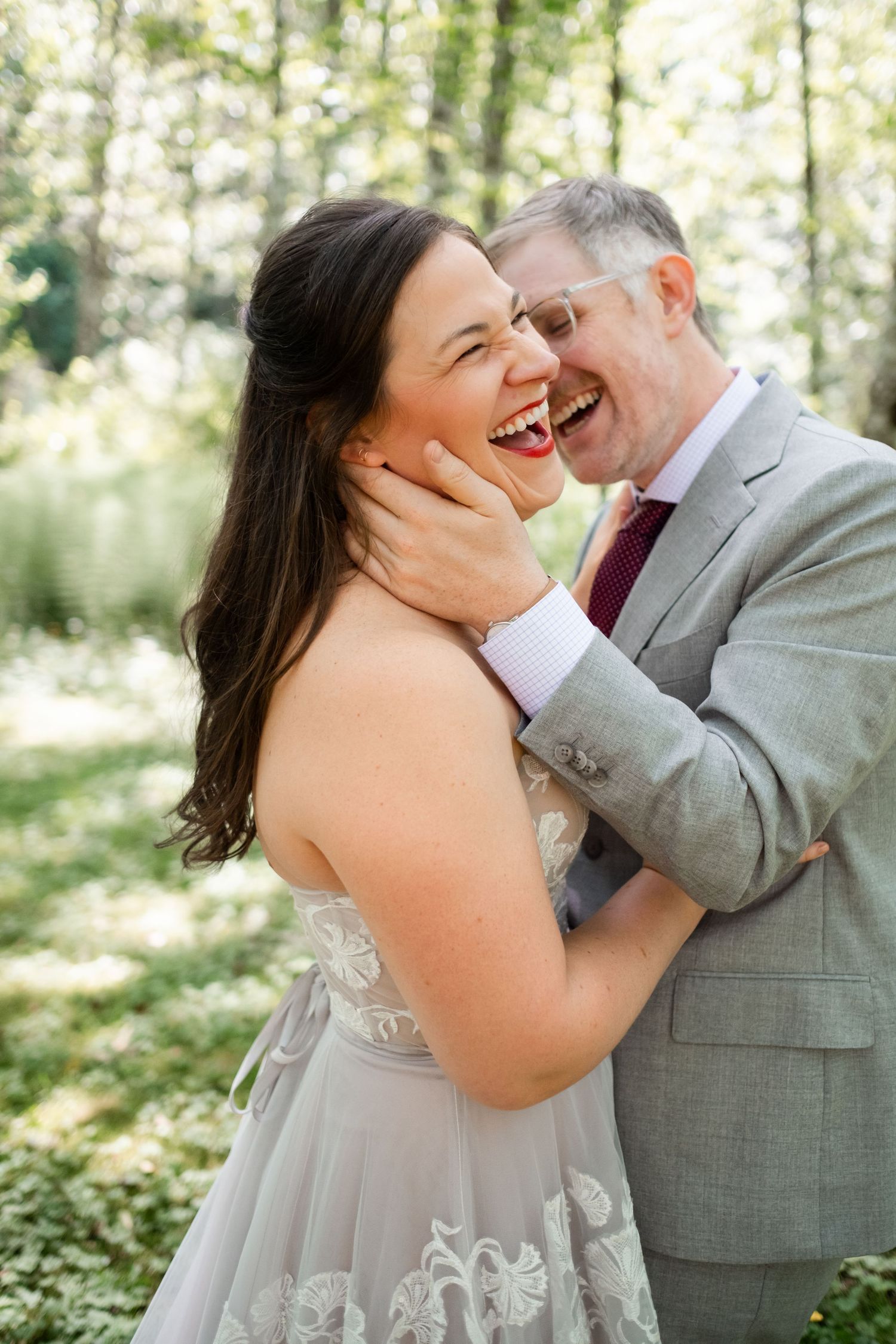Joyful bride and groom embracing outdoors, laughing together in a sunlit forest setting on their wedding day.