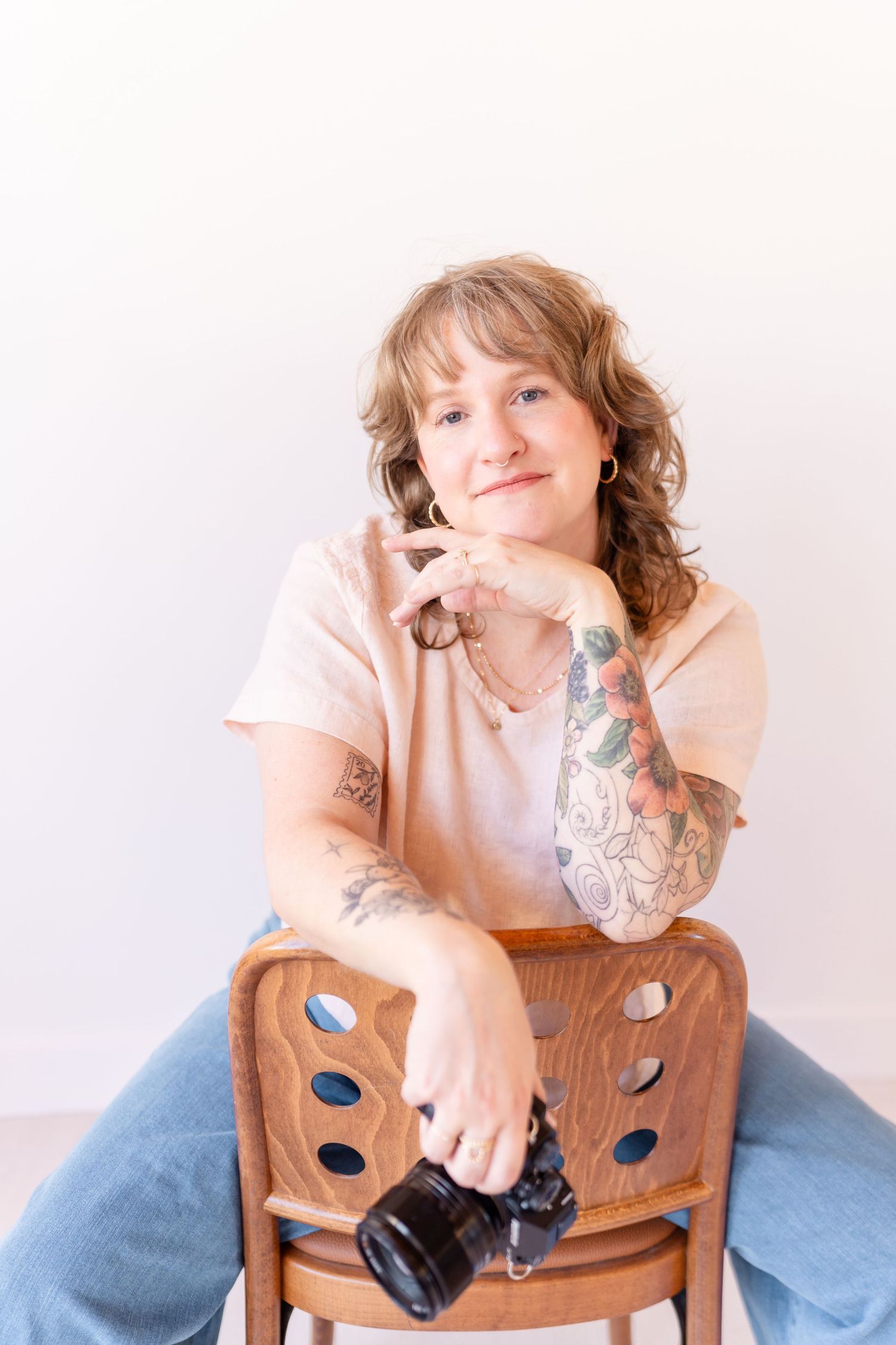 Tattooed woman sitting on wooden chair holding camera, wearing pink tee and jeans, smiling against white background.