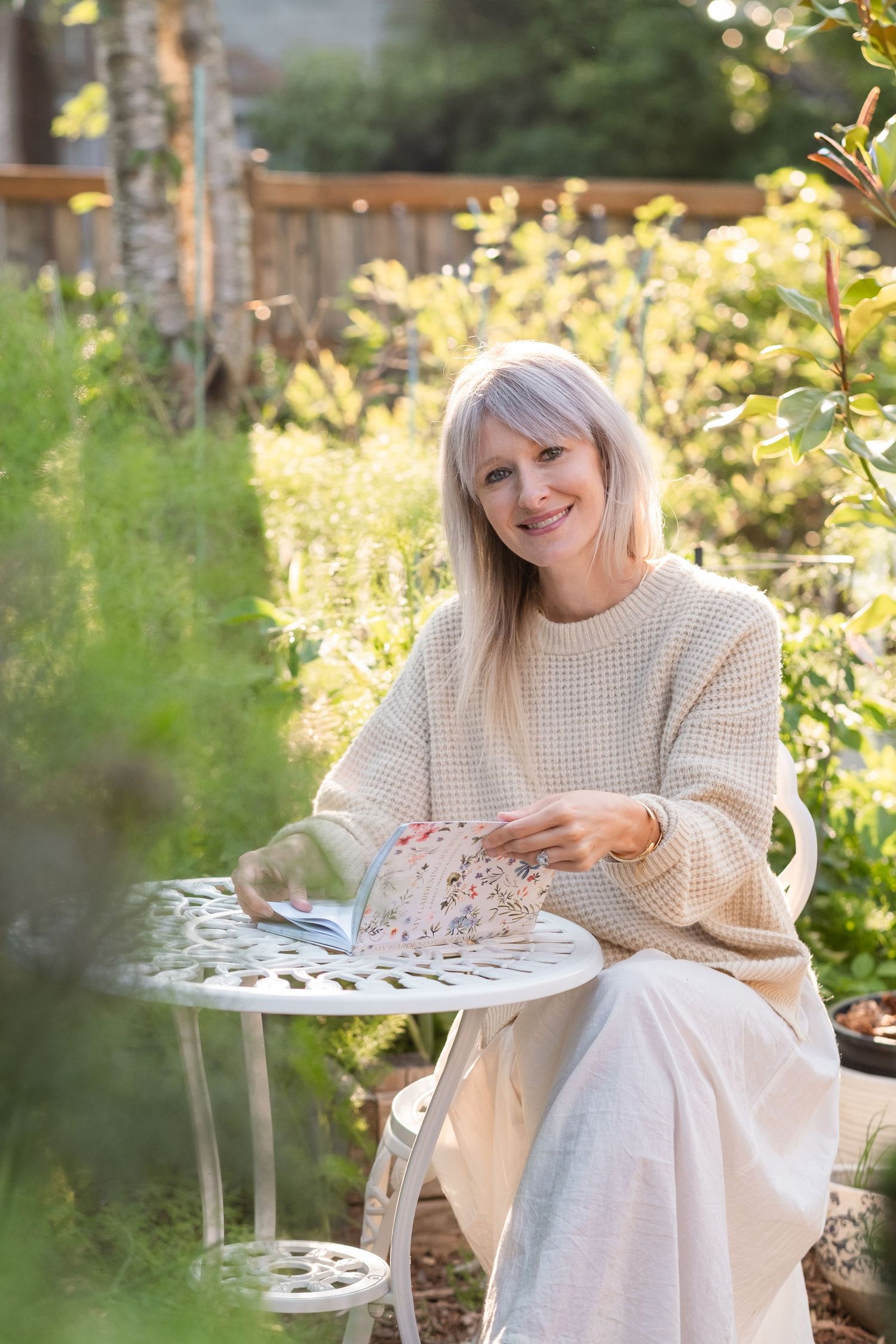 Smiling blonde woman in beige sweater holding a floral book at a white garden table.