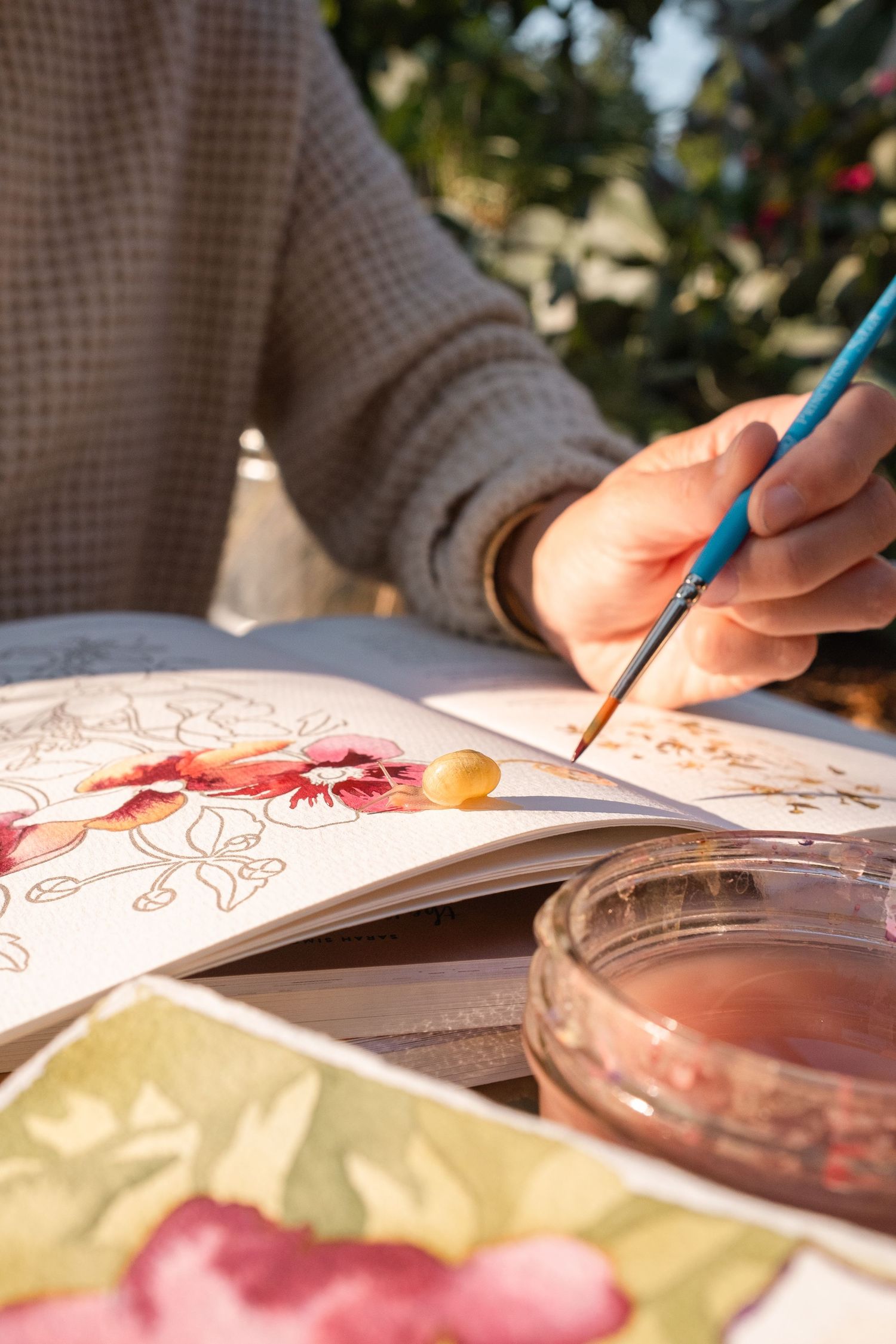 Close-up of hands painting watercolor flowers in a sketchbook outdoors with a blue brush.