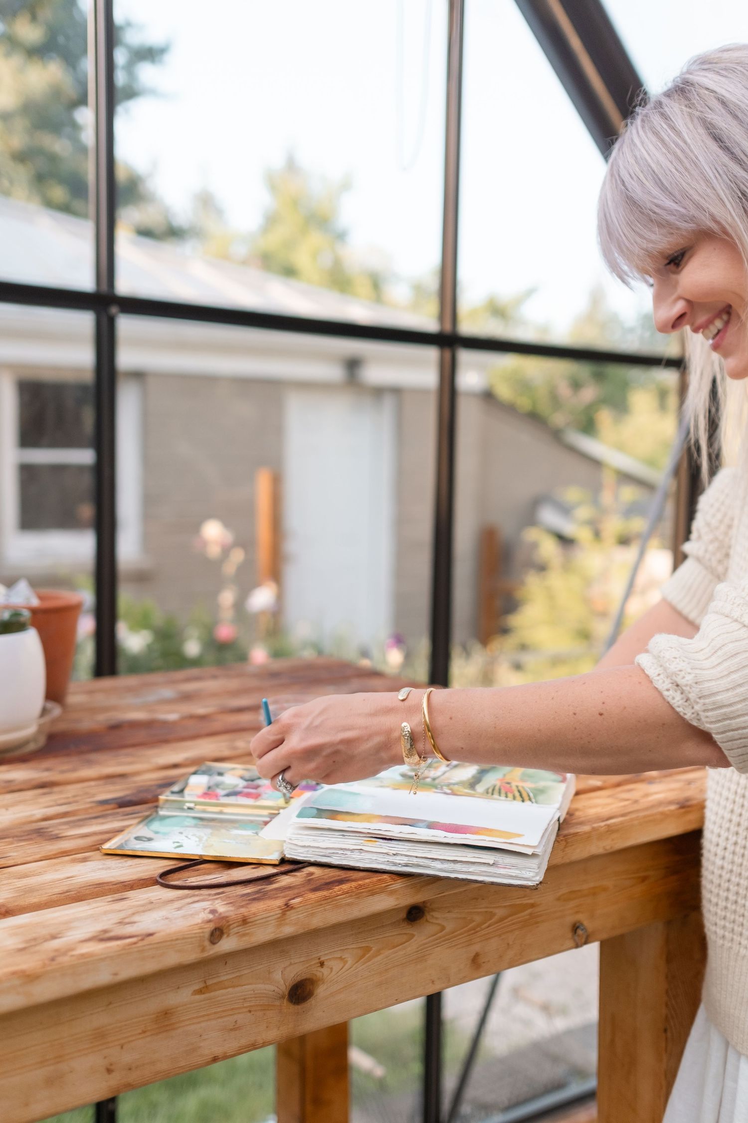 Smiling woman with lavender hair writing in a sketchbook at a wooden table in a greenhouse.