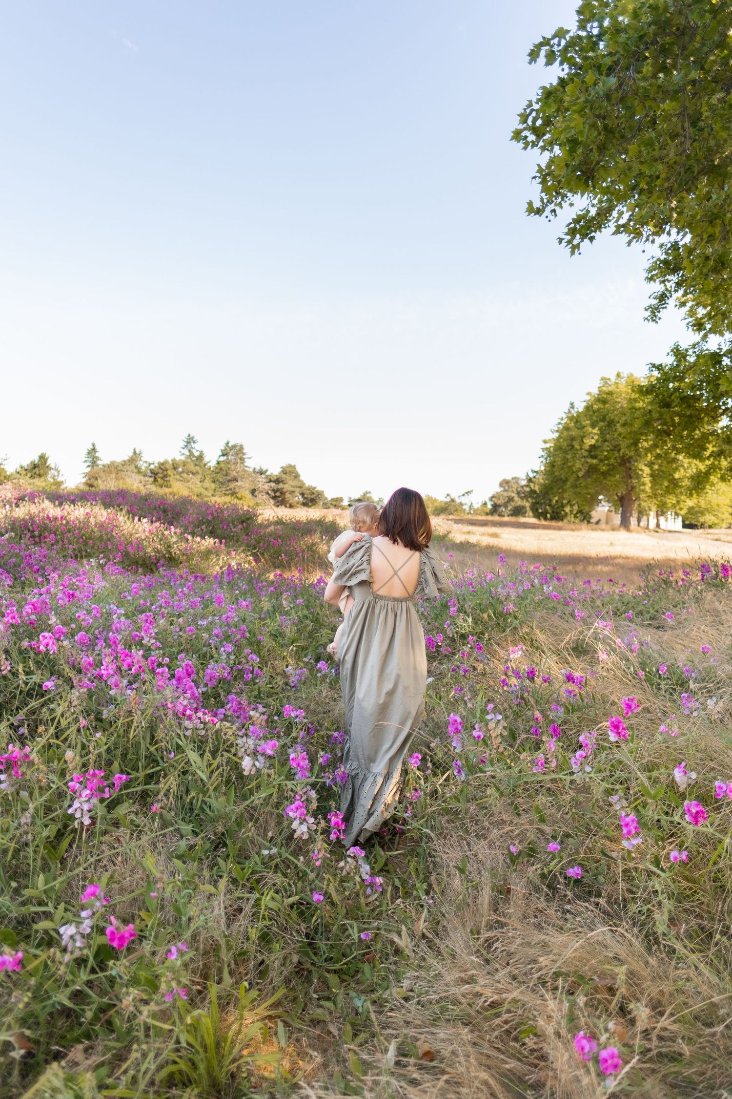 A person in a flowing taupe dress walks through a meadow of vibrant purple wildflowers at sunset.