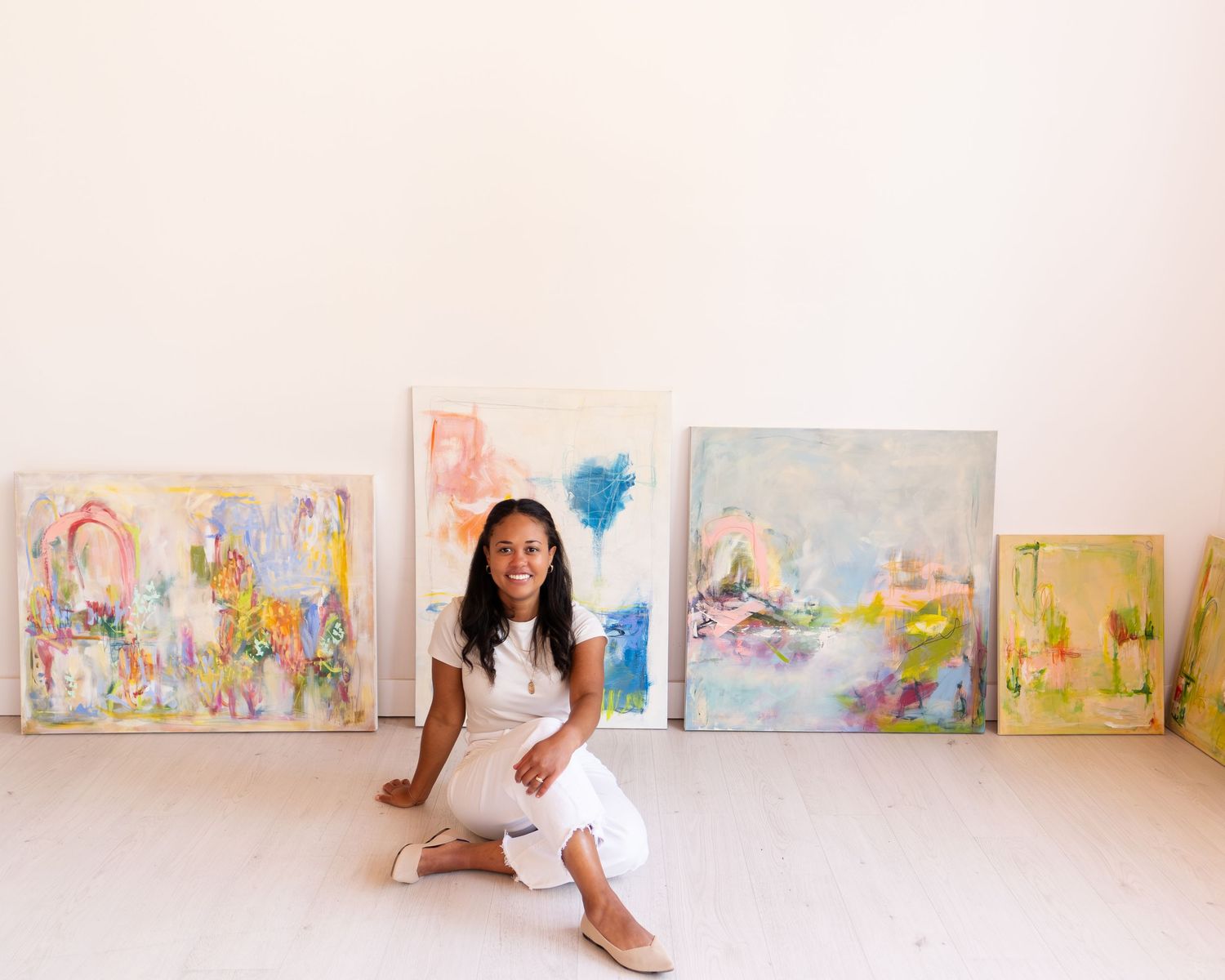 Artist sitting on floor in white outfit surrounded by colorful abstract paintings in a bright studio.