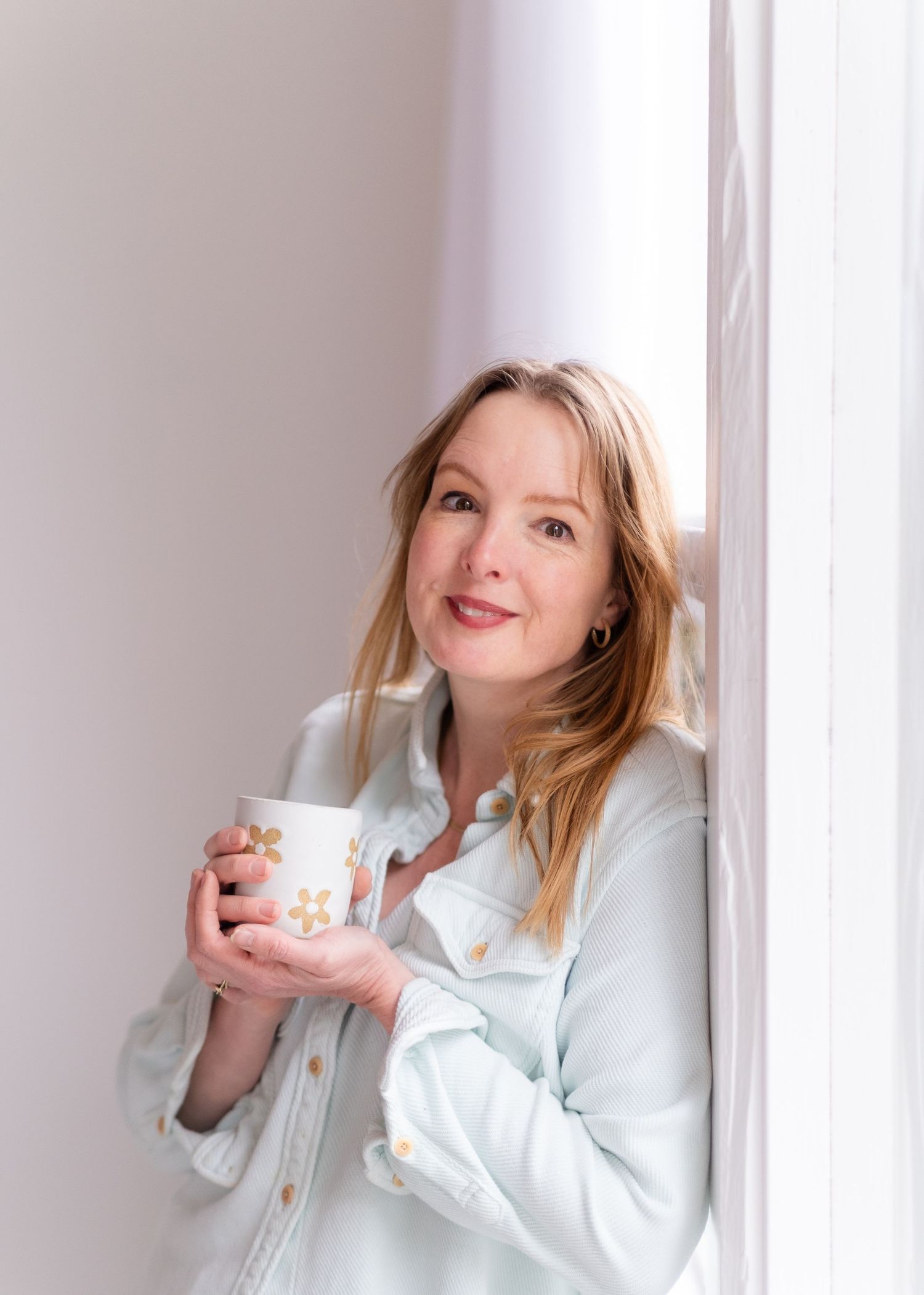 Person in light blue button-down shirt holding a ceramic mug while leaning against white wall in bright setting.