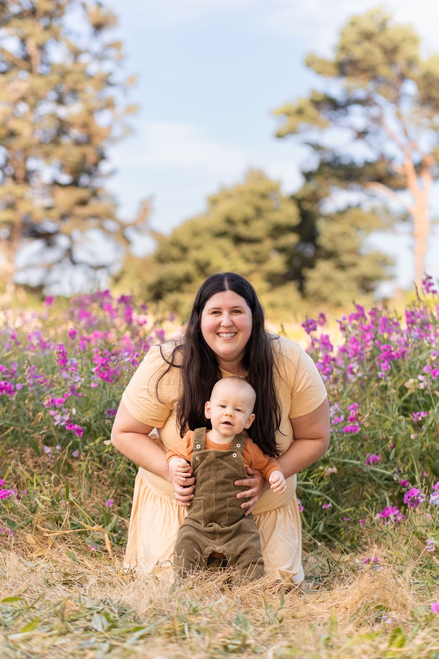 A person in a yellow dress kneels with a small child wearing brown overalls in a field of purple wildflowers.