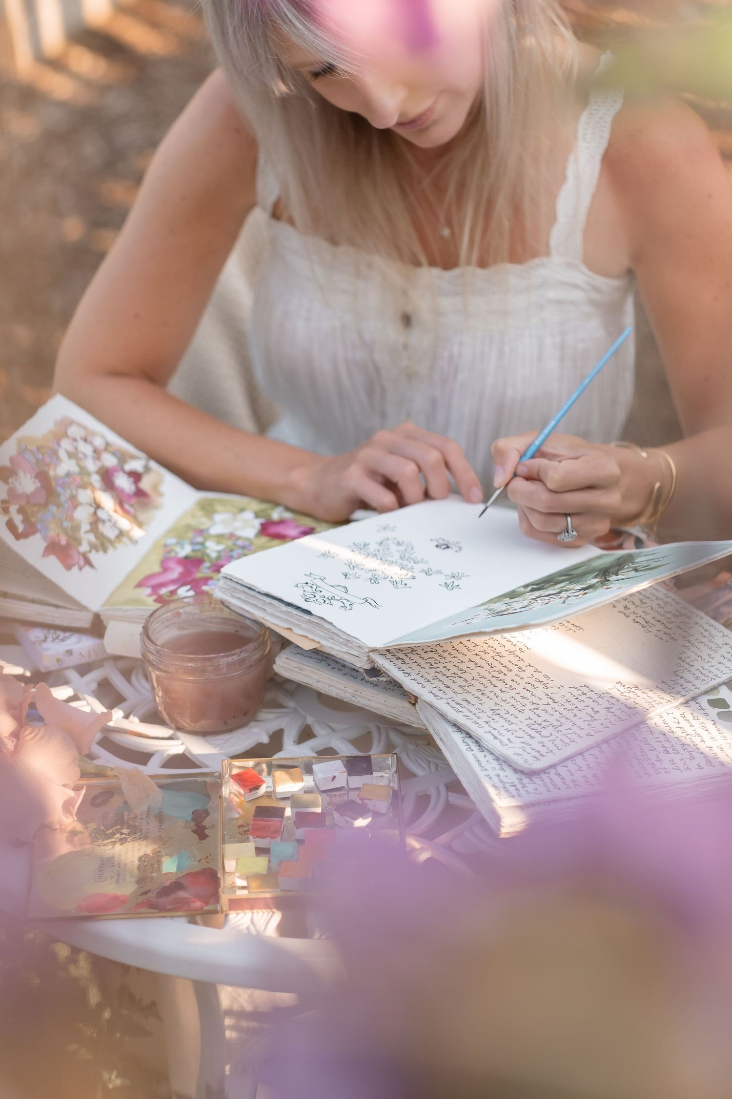 Blonde woman painting watercolor florals in a sketchbook at an outdoor garden table.